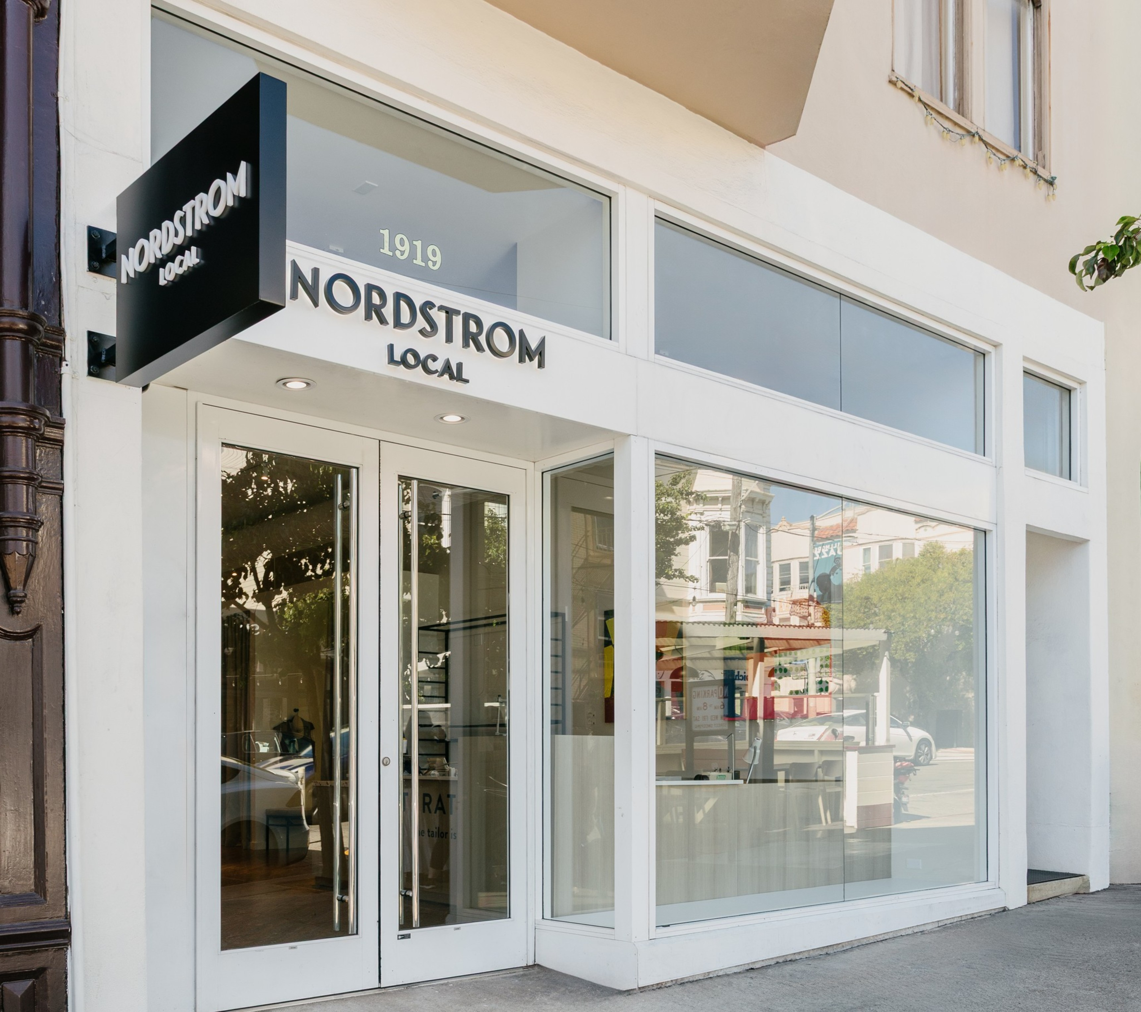 A Nordstrom Local store with glass doors and large windows, displaying reflections of nearby buildings and trees on a city street.
