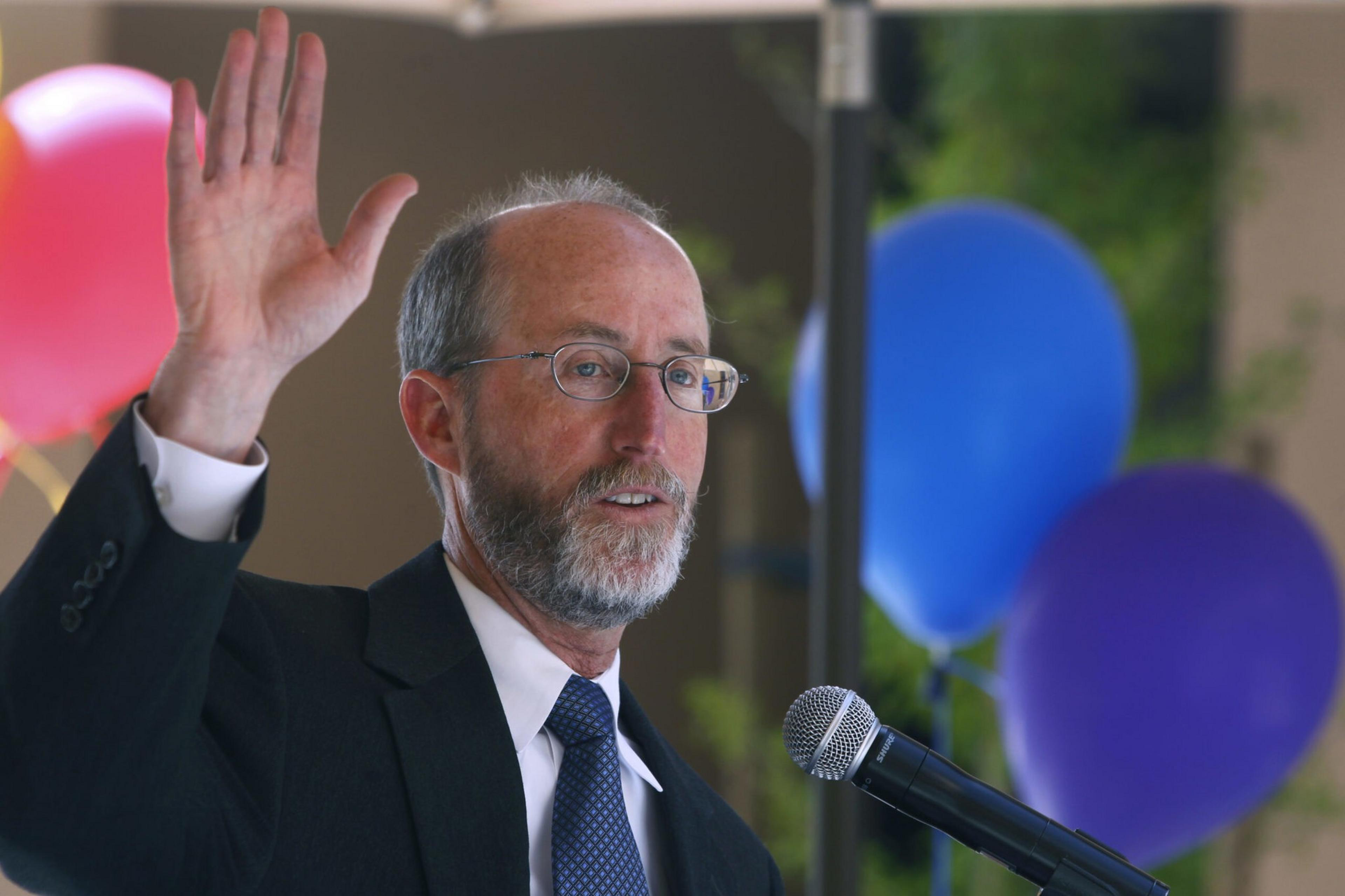 A man with glasses and a beard in a suit raises his hand while speaking into a microphone, with colorful balloons in the background.