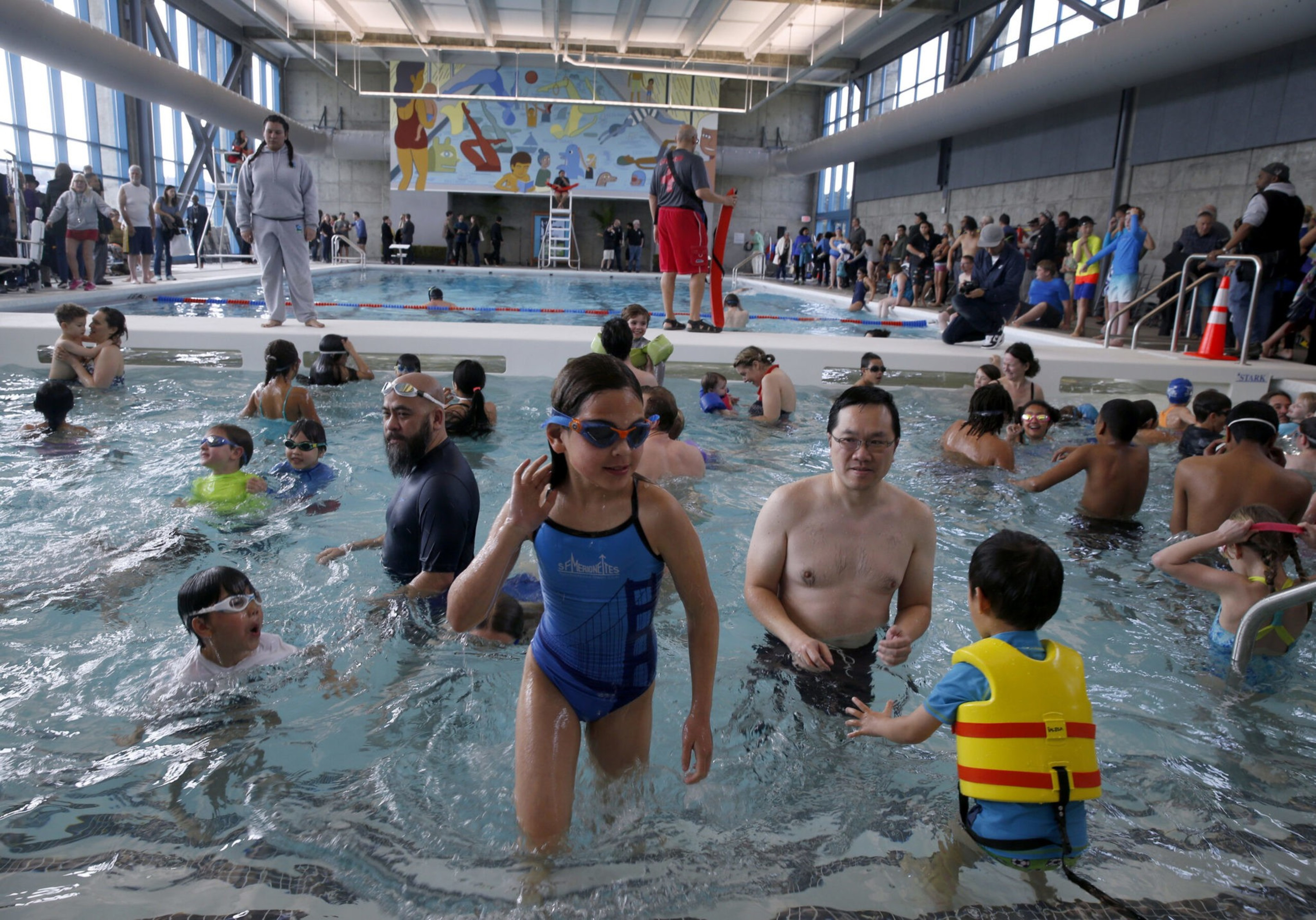 A crowded indoor swimming pool with children and adults swimming, wearing goggles and swimwear, while others watch from the poolside and a lifeguard stands nearby.