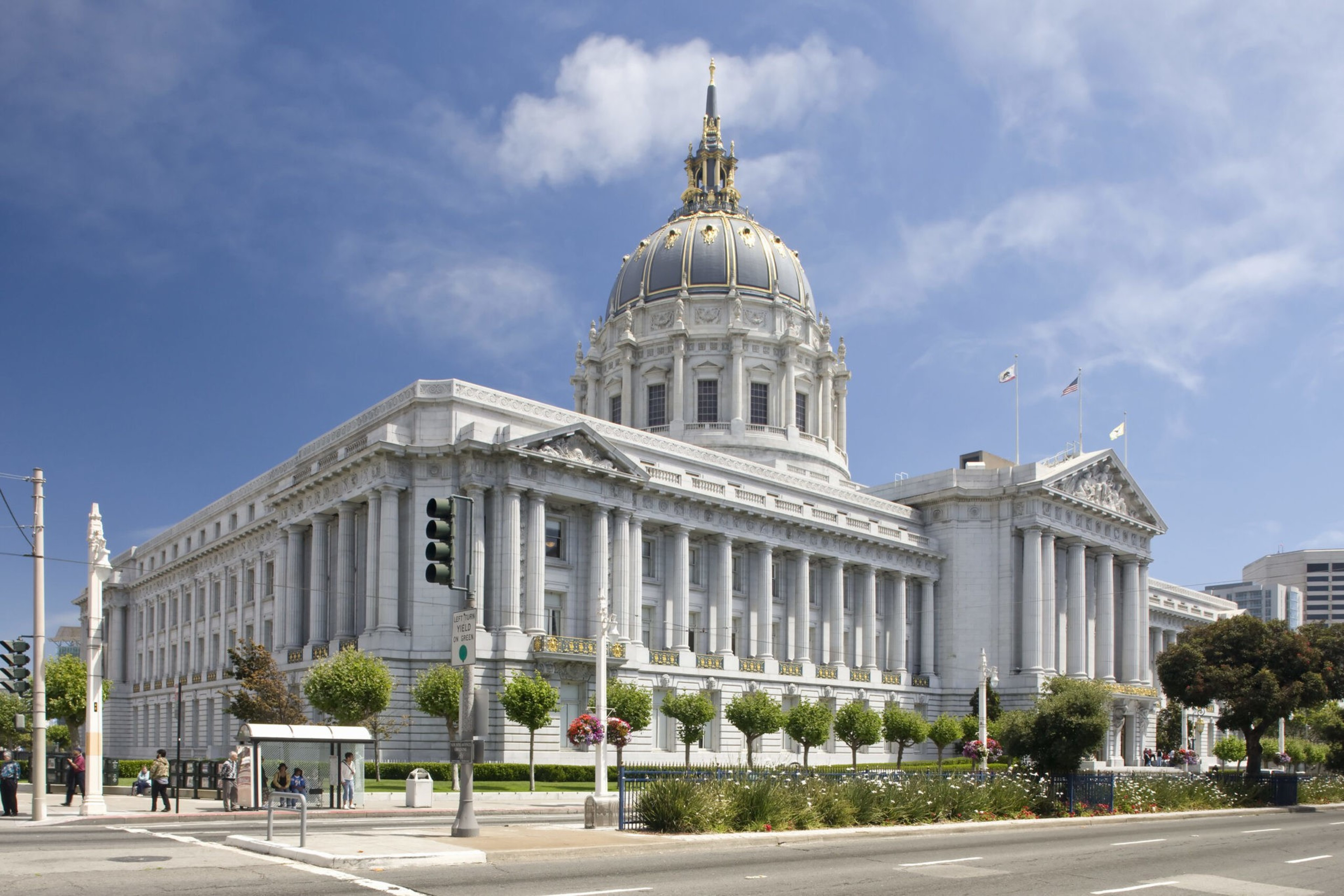 A grand neoclassical building with a large domed roof adorned with gold accents sits on a sunny street corner, surrounded by neatly trimmed trees and flowers.