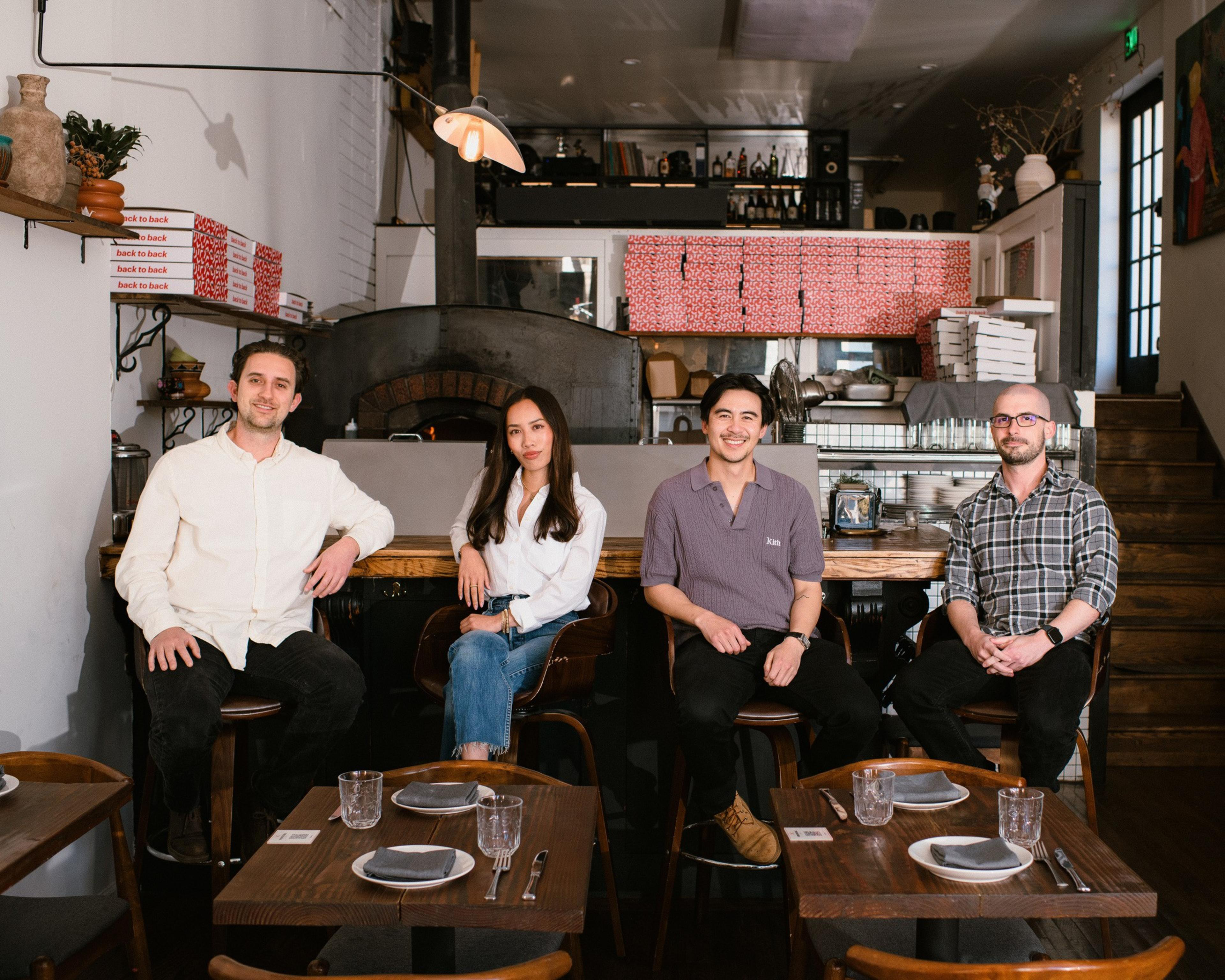 Four people are seated at a cozy restaurant counter. They smile towards the camera. The background includes stacked pizza boxes and a wood-fired oven.