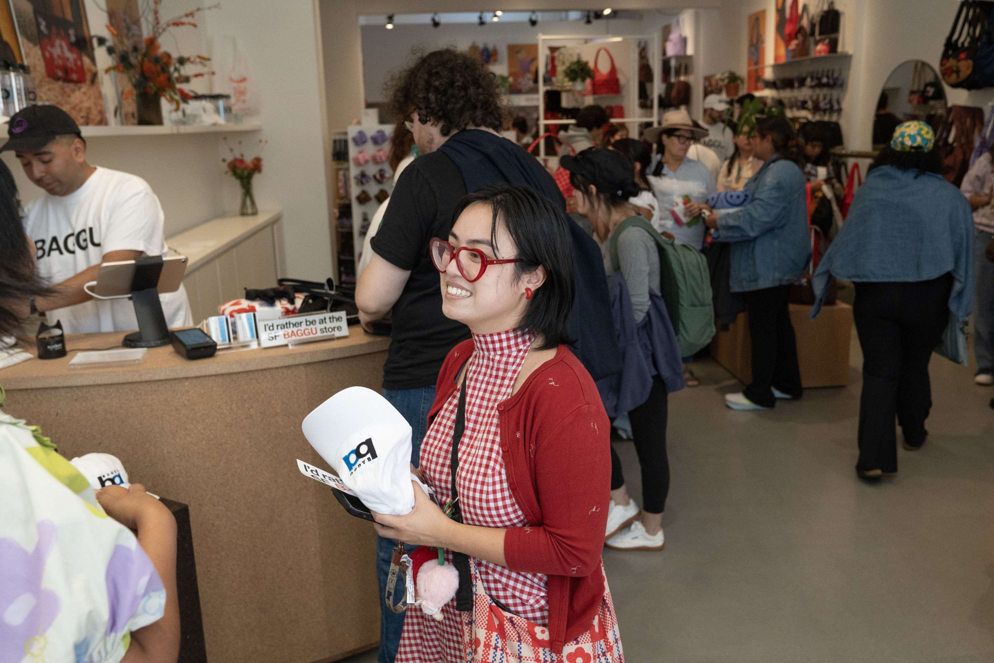 A smiling woman in red glasses and a red checkered dress holds a white cap in a busy store with other shoppers and colorful bags on display.
