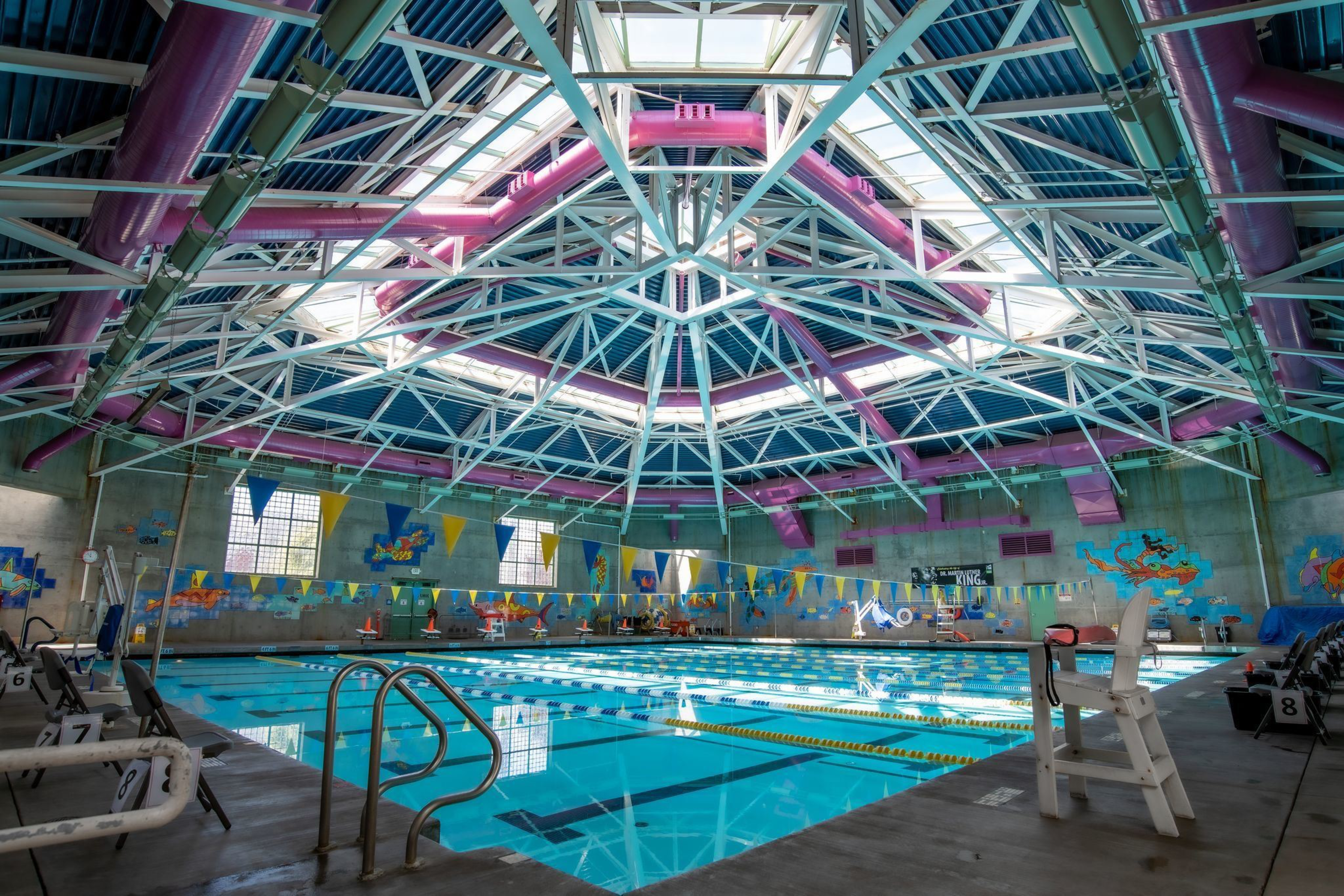 An indoor swimming pool with lane dividers, colorful banners, purple ductwork on the ceiling, and murals painted on concrete walls.