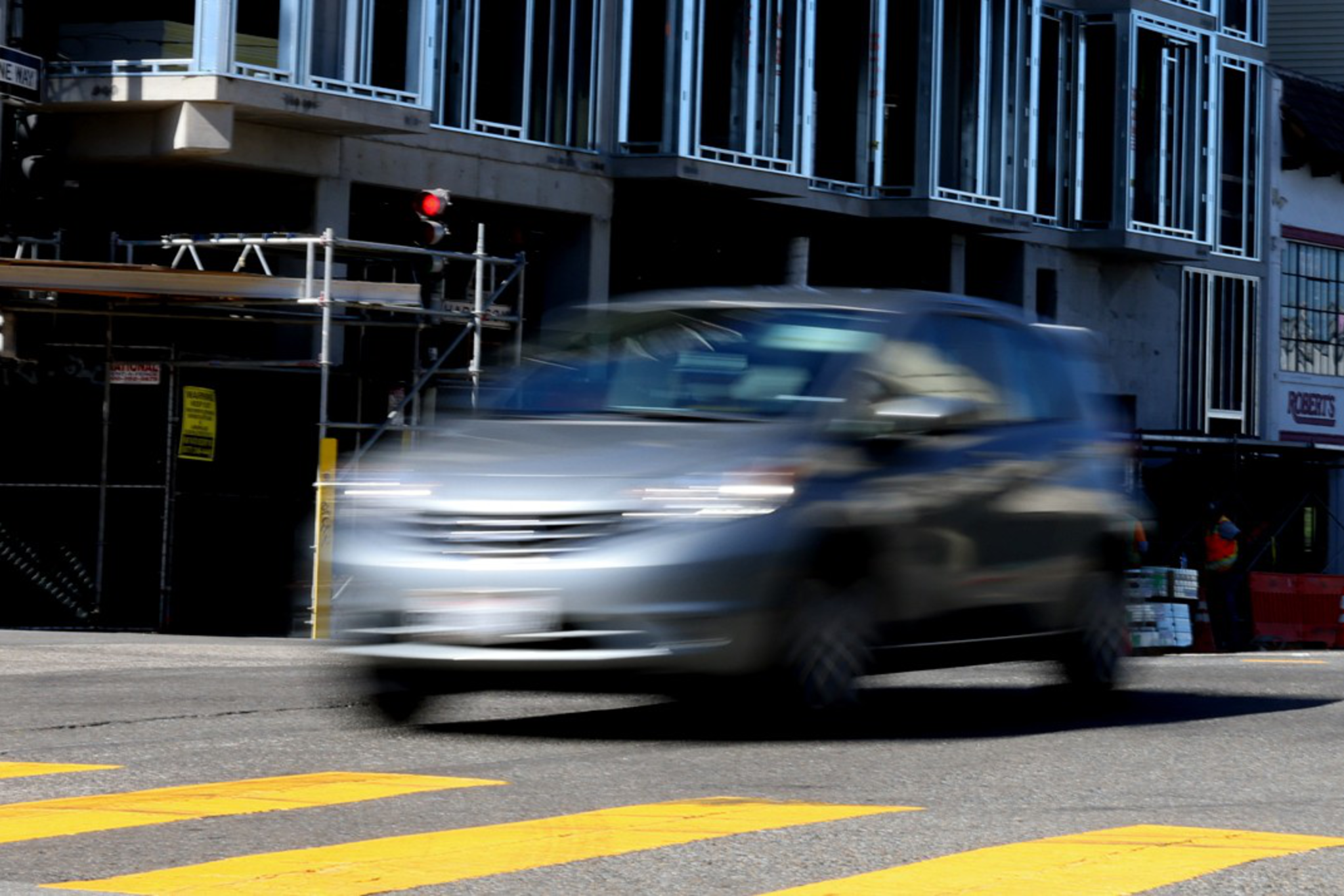 A silver car is speeding past a yellow pedestrian crosswalk in front of a building under construction with scaffolding.