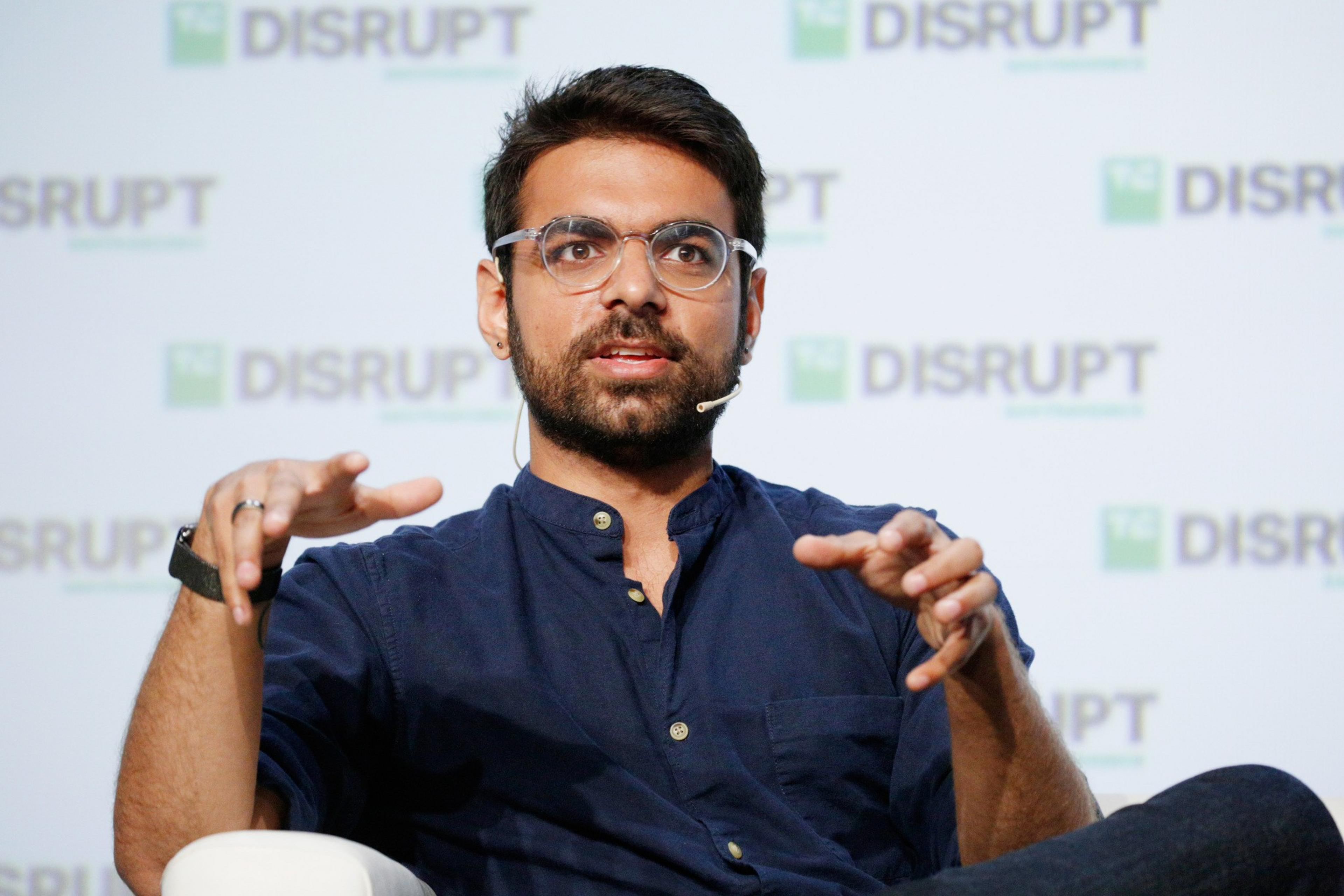 A man with glasses and a beard speaks animatedly, wearing a dark blue shirt and a headset microphone. The backdrop features the word &quot;DISRUPT&quot; repeatedly.