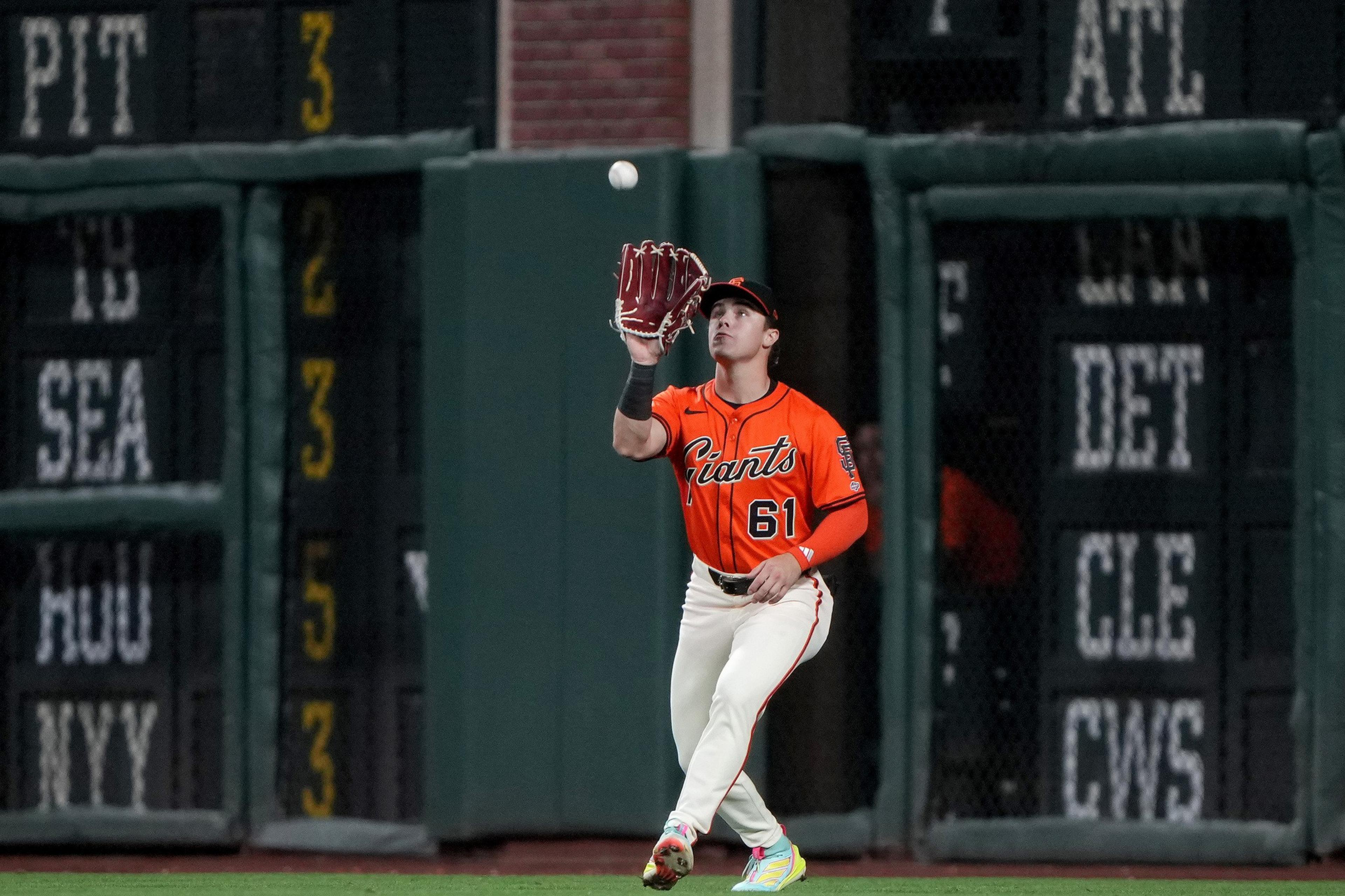 A baseball player in an orange Giants jersey with number 61 is preparing to catch a ball while standing on the field near the outfield wall.