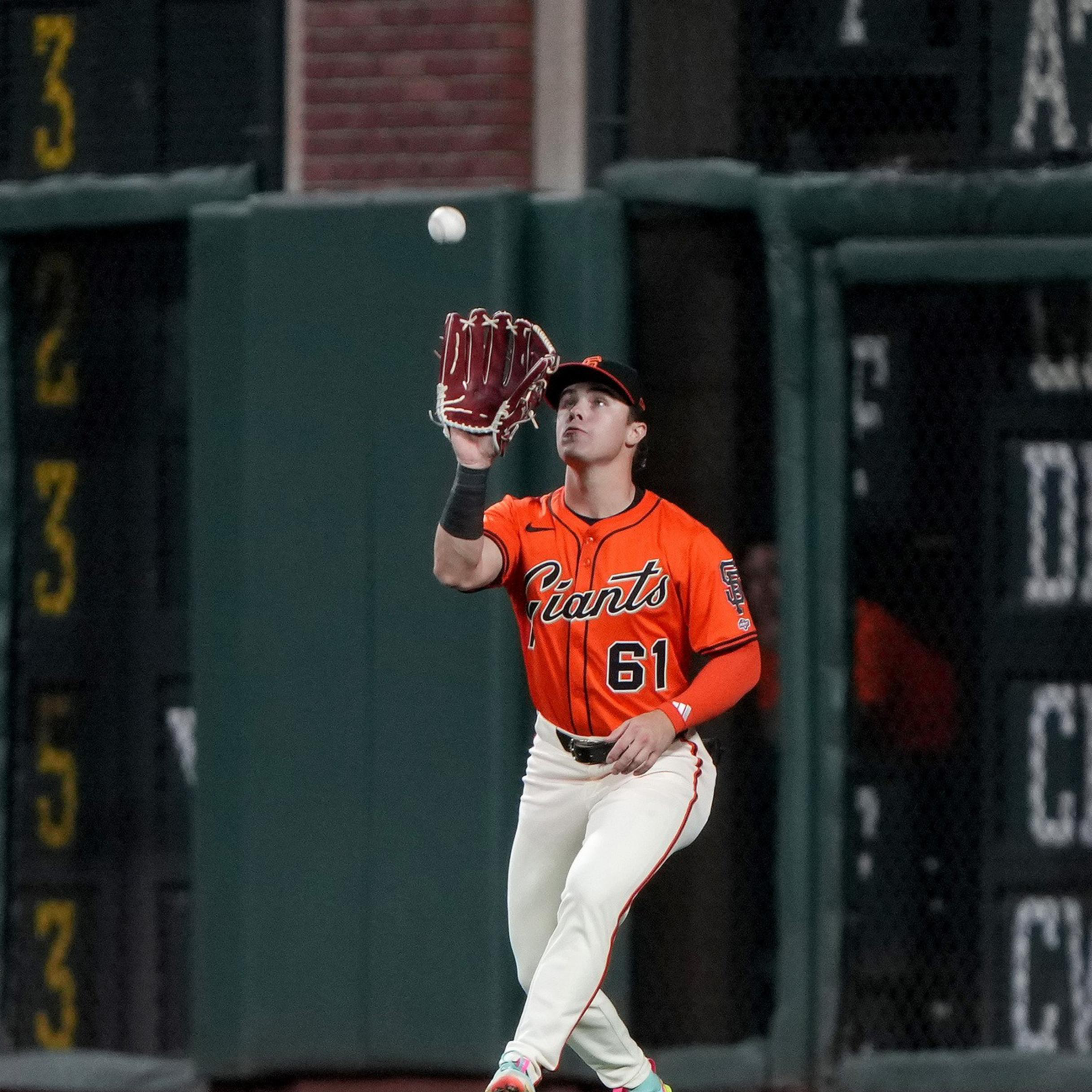A baseball player in an orange Giants jersey with number 61 is preparing to catch a ball while standing on the field near the outfield wall.