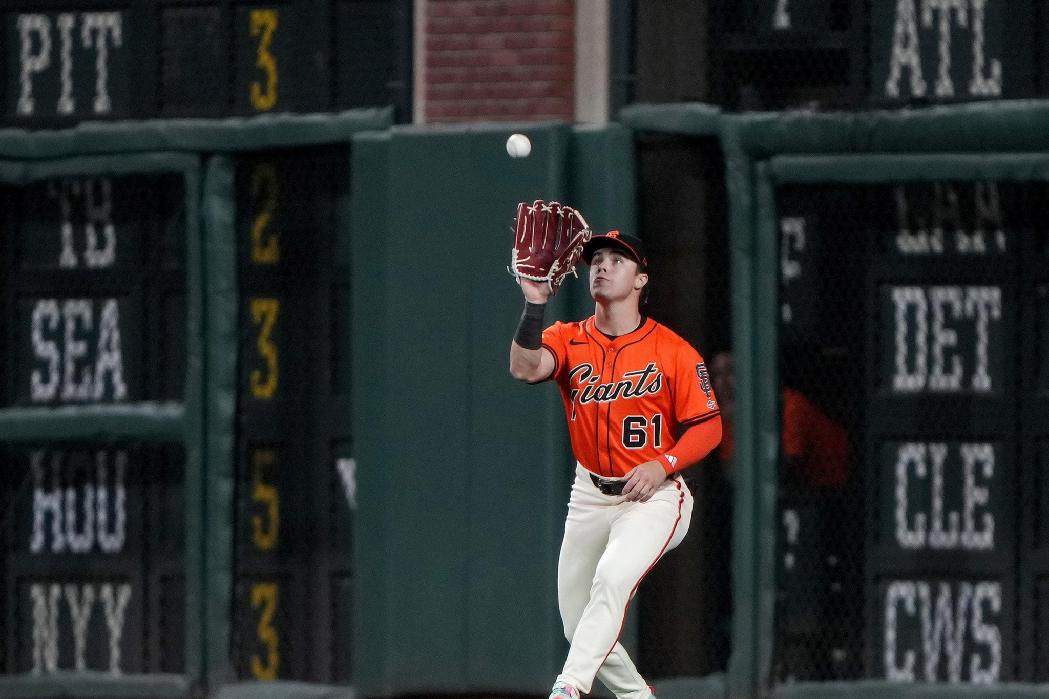 A baseball player in an orange Giants jersey with number 61 is preparing to catch a ball while standing on the field near the outfield wall.