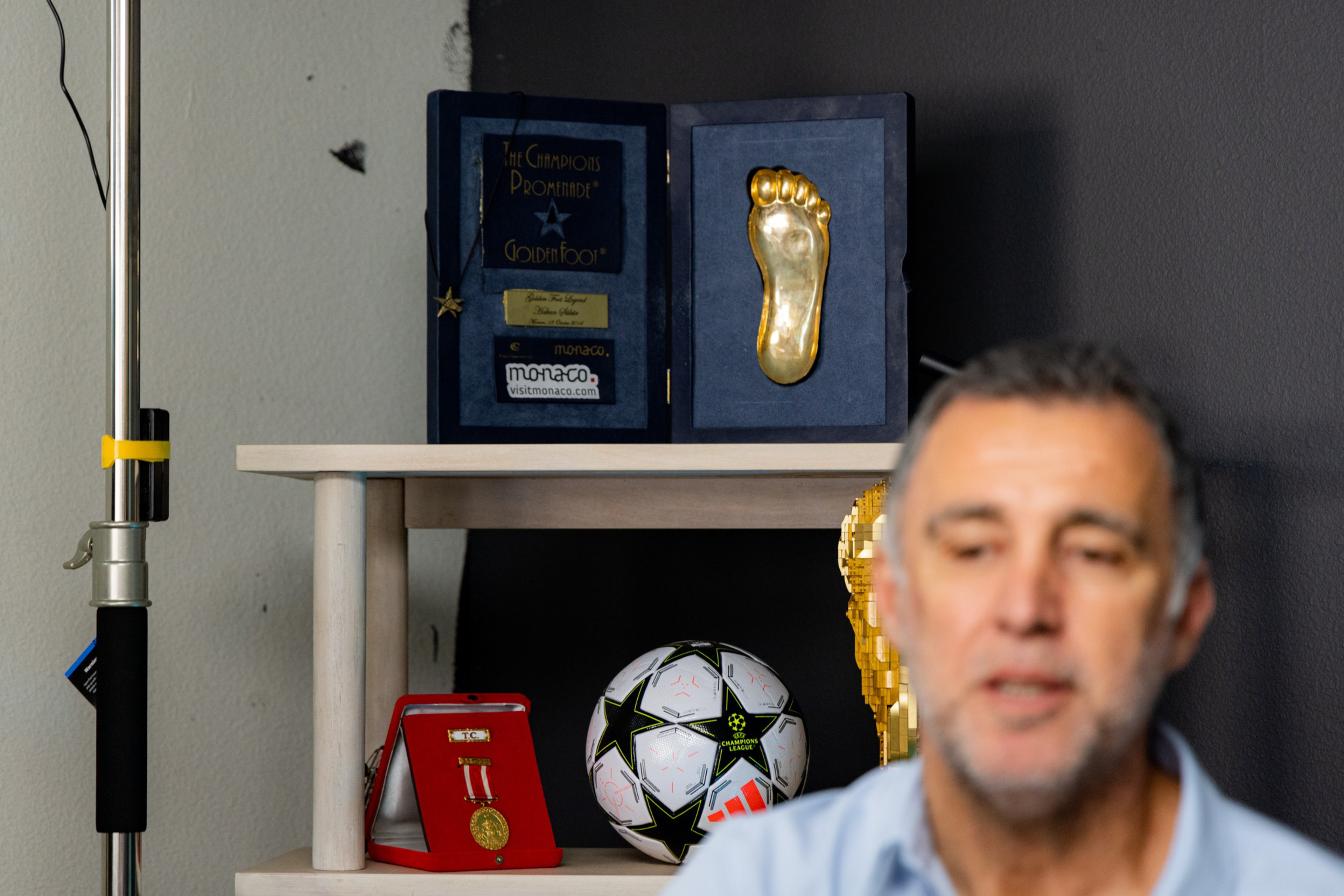 A golden foot award, a framed medal, a Champions League soccer ball, and a golden trophy are displayed on a shelf behind a man.