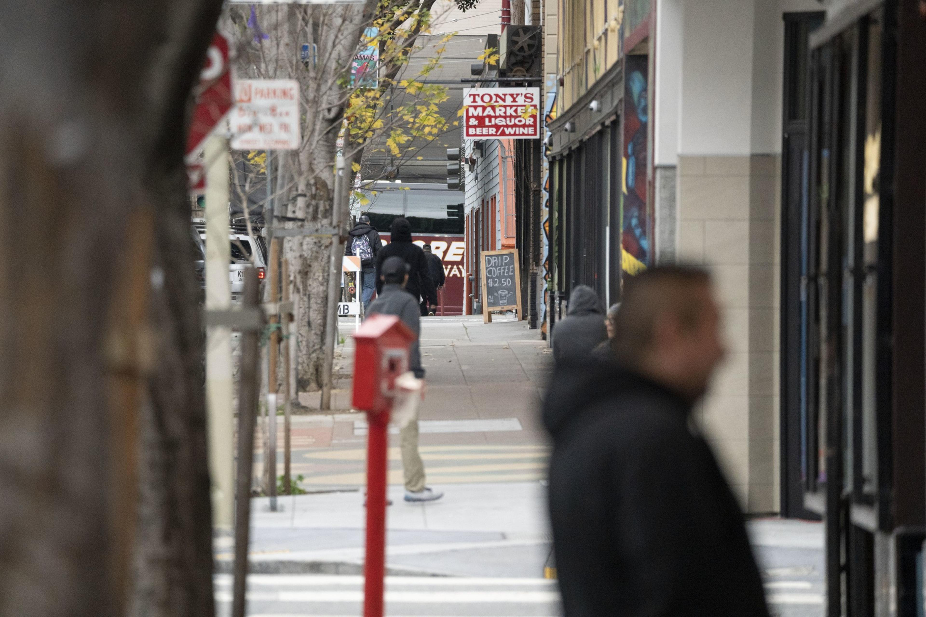 A bustling city sidewalk is lined with trees, people in jackets walk past shops. A &quot;Tony's Market &amp; Liquor&quot; sign is visible, with a board for drip coffee.