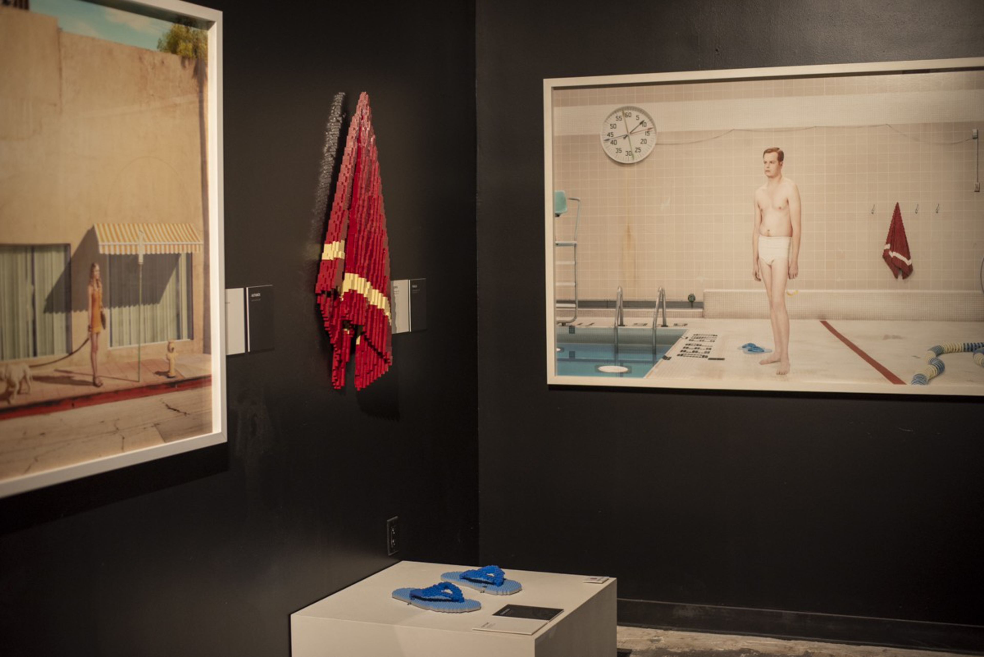 A gallery corner displays two large framed photos and a red towel made of blocks on a black wall, with a white pedestal holding blue block sculptures.