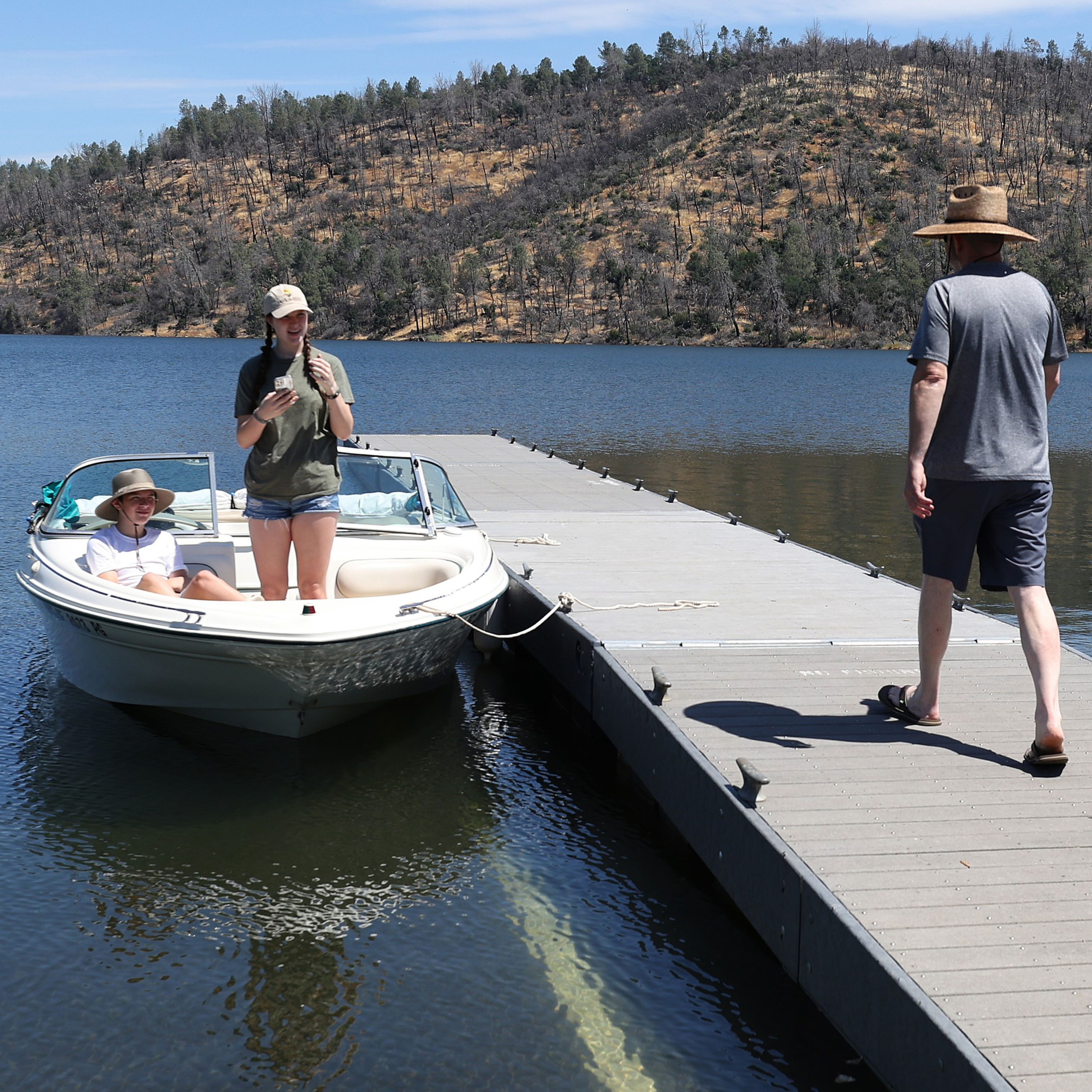 A boat with two people onboard is docked beside a pier. Another person is walking away on the dock. The scene is set on a calm lake with a hilly, forested background.