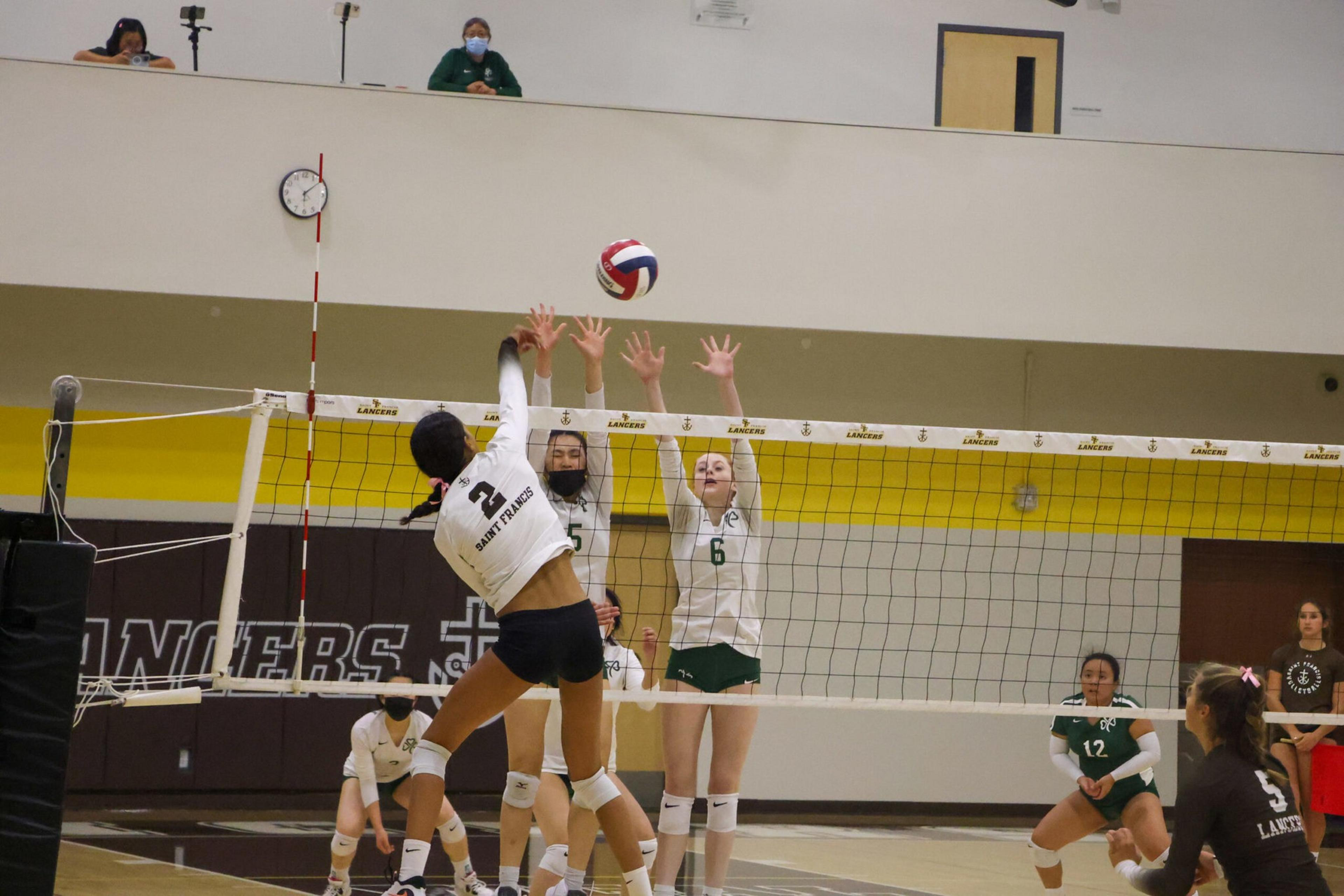 A volleyball player from Saint Francis jumps to spike the ball while two Lancers players jump to block it at the net during a game.