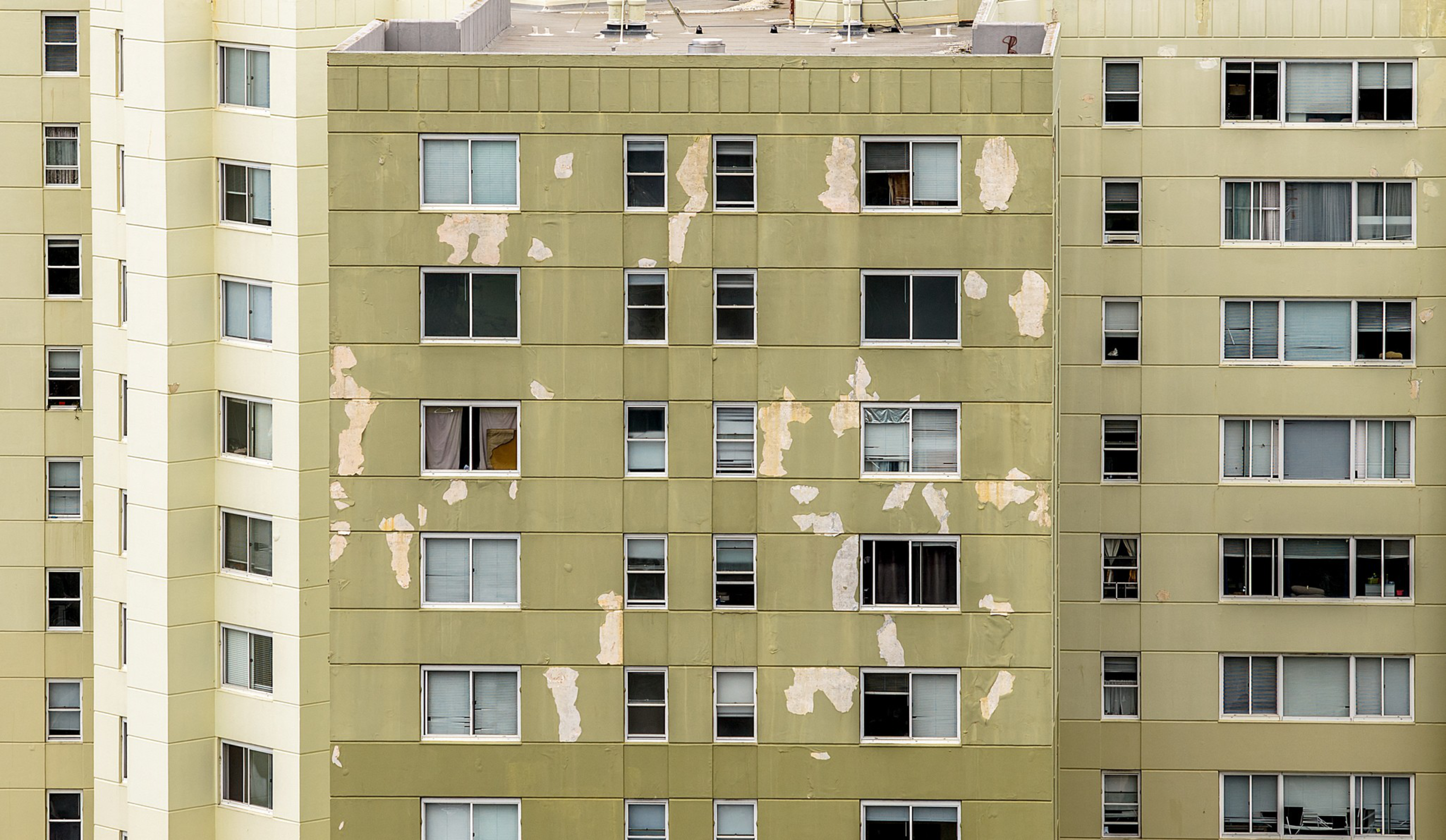 A beige apartment building with many windows, some open and some closed, with peeling paint visible in multiple patches across the facade.
