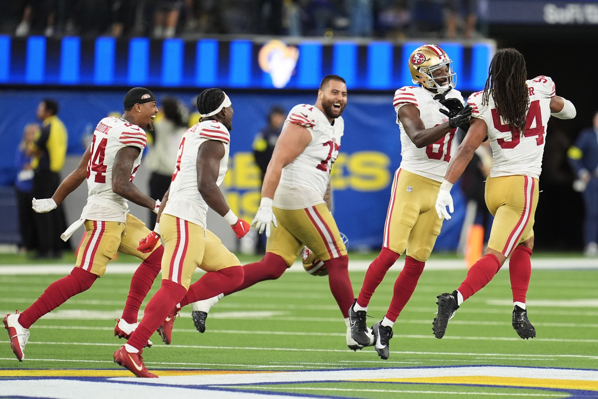 Five San Francisco 49ers players in white and gold uniforms celebrate enthusiastically on a football field with a blue and yellow background.