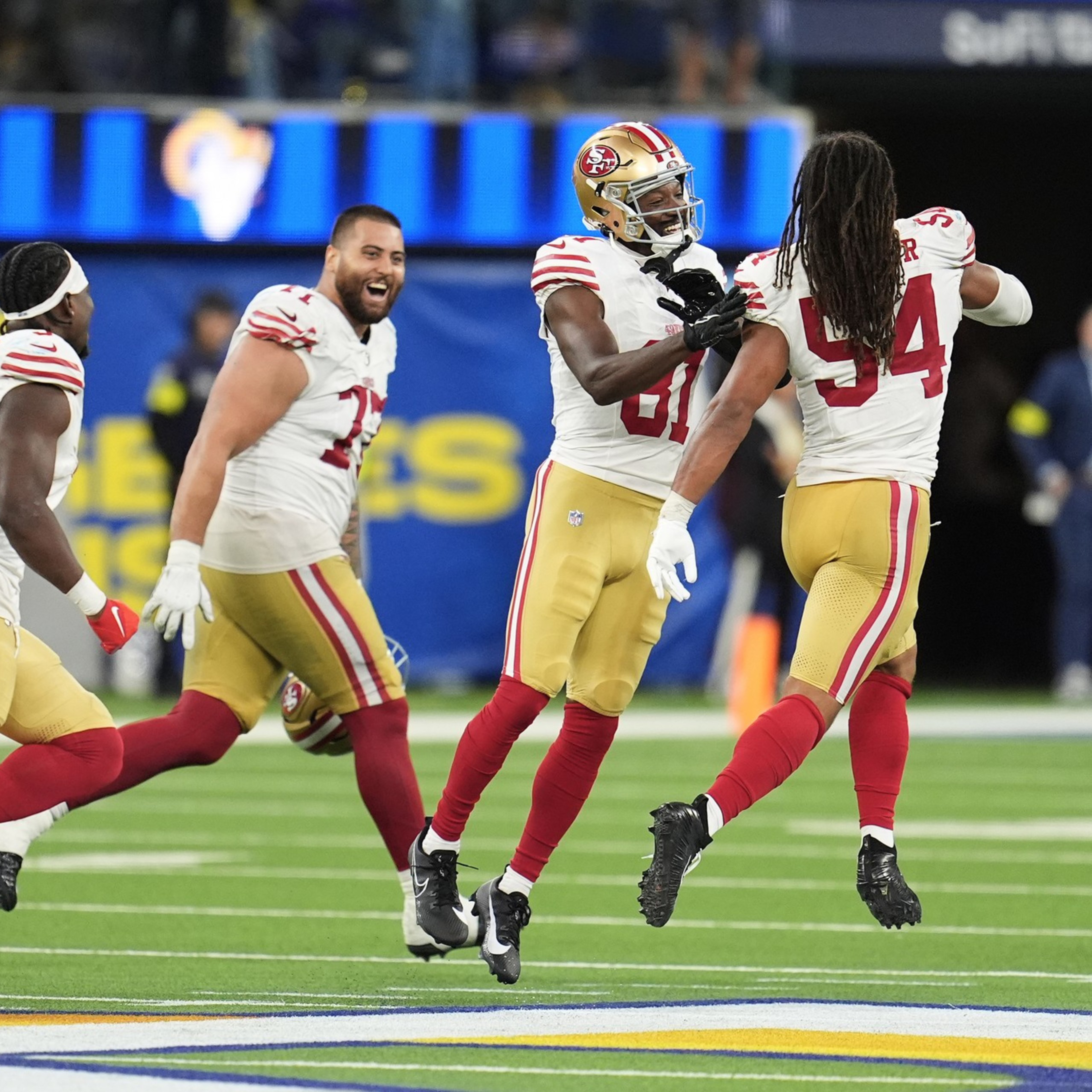 Five San Francisco 49ers players in white and gold uniforms celebrate enthusiastically on a football field with a blue and yellow background.