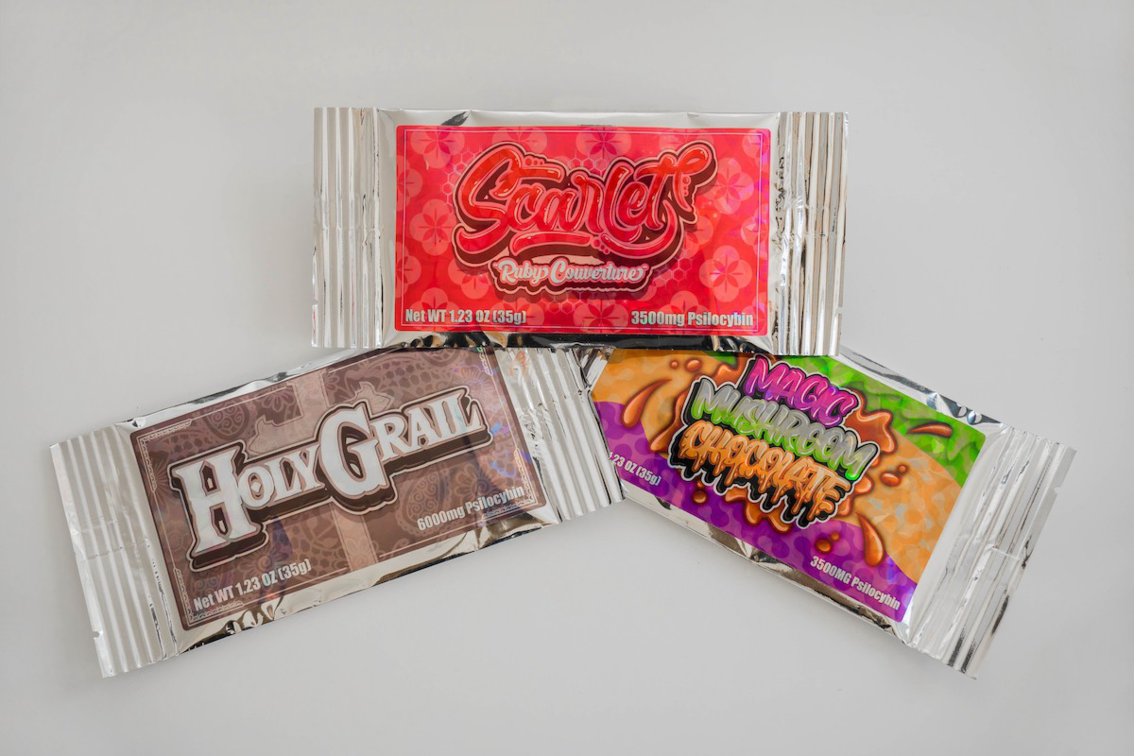 Three packaged edibles with colorful labels named Scarlet Ruby Convertine, Holy Grail, and Magic Mushroom Grapple, each containing psilocybin.