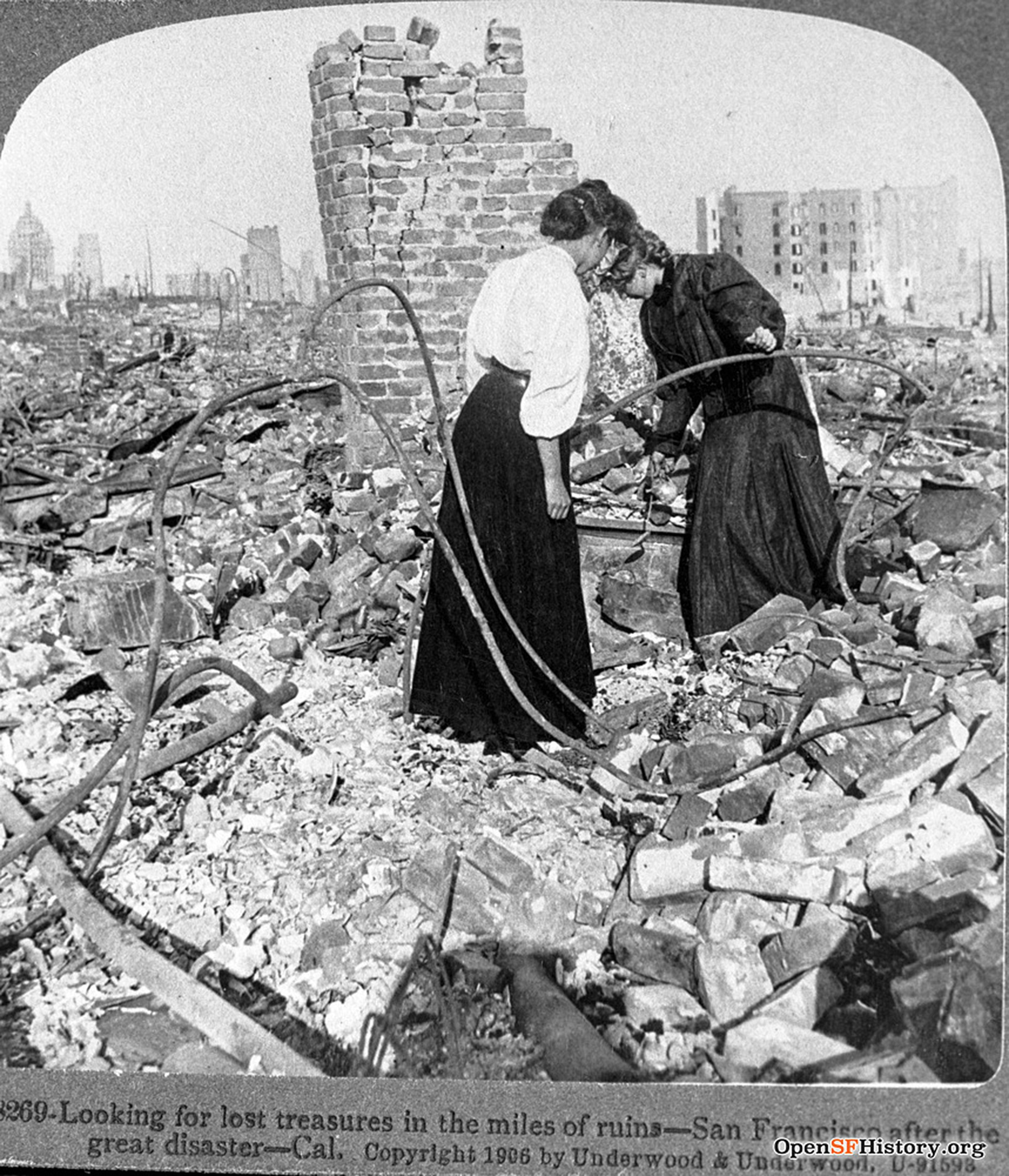Two women in long skirts search through rubble and debris near a partially standing brick chimney in a city devastated by disaster.