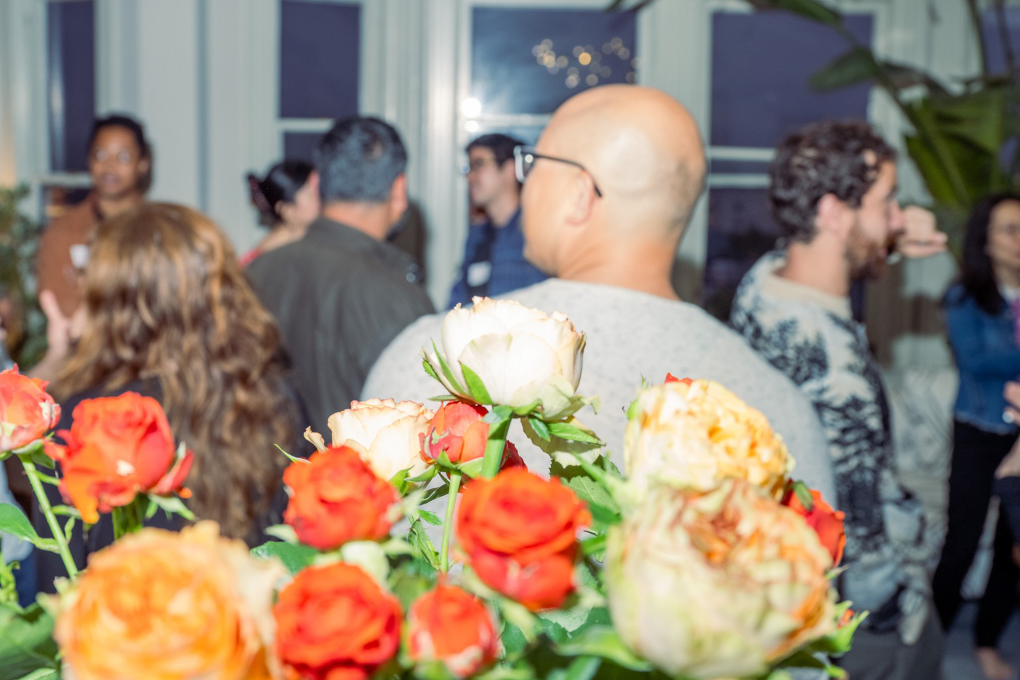 A bouquet of orange and white roses is in focus with a group of people socializing in a warmly lit room with windows and a modern chandelier.