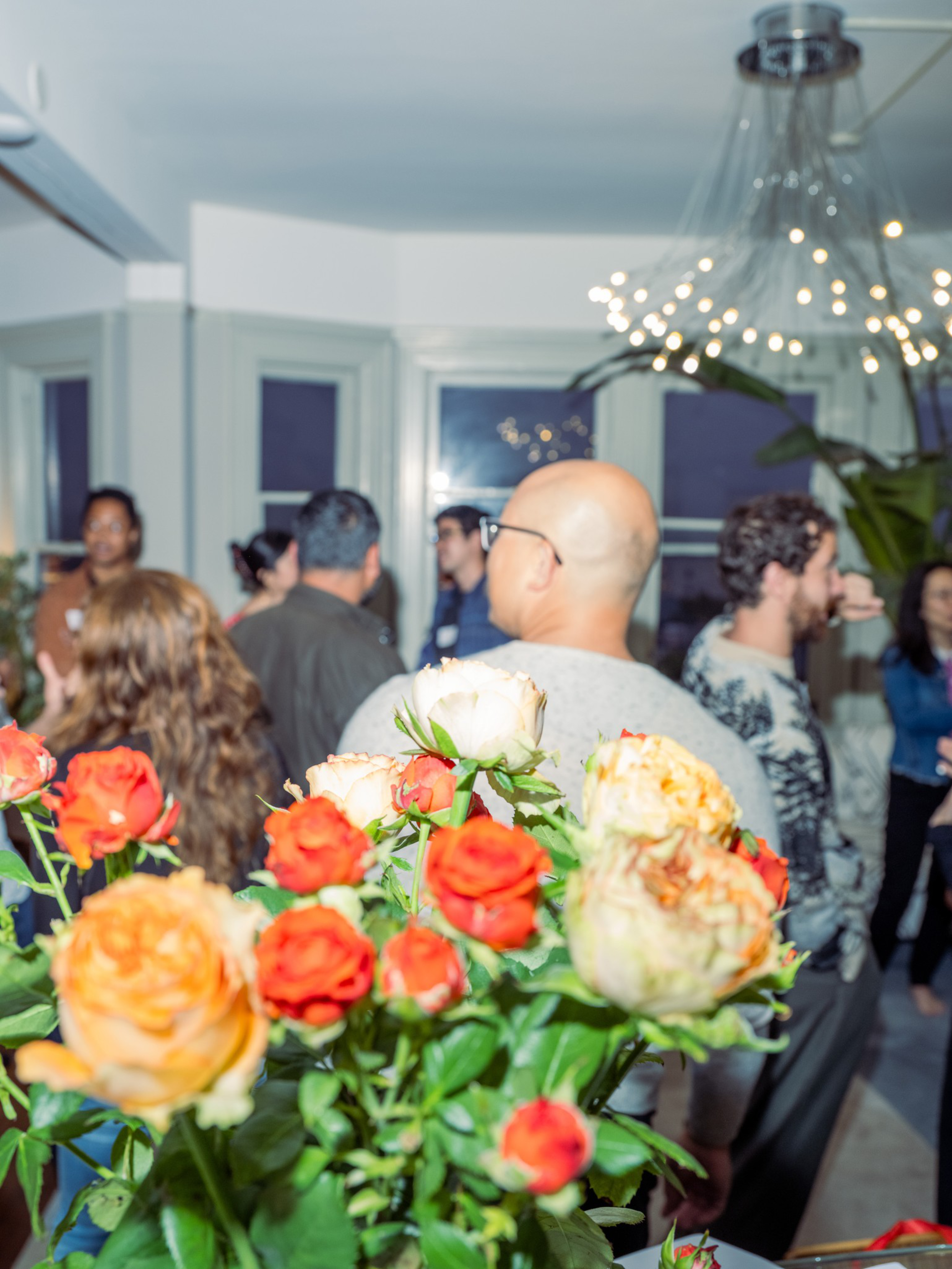 A bouquet of orange and white roses is in focus with a group of people socializing in a warmly lit room with windows and a modern chandelier.