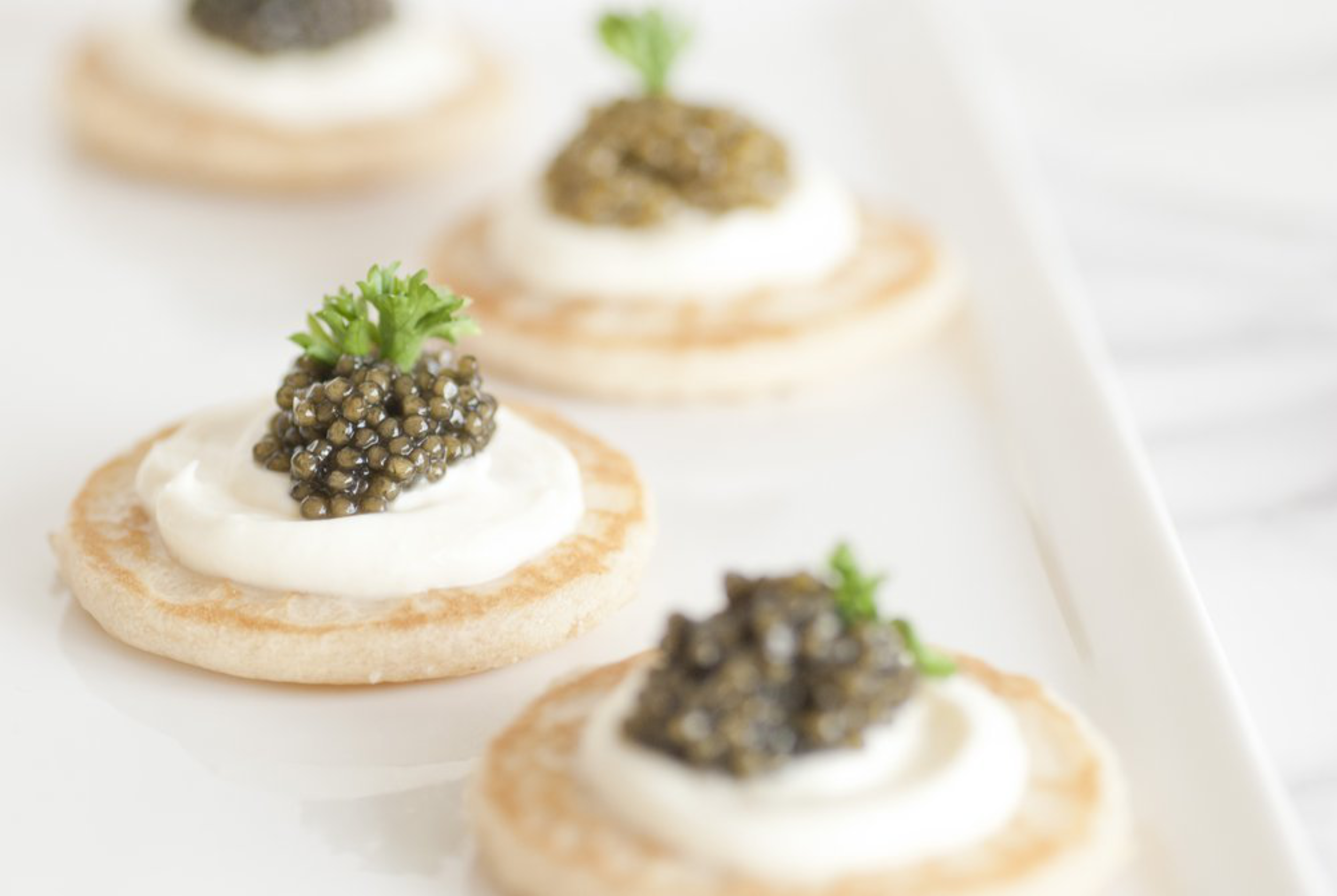 Mini blinis topped with a dollop of cream, a serving of caviar, and a small sprig of parsley, arranged on a white plate.