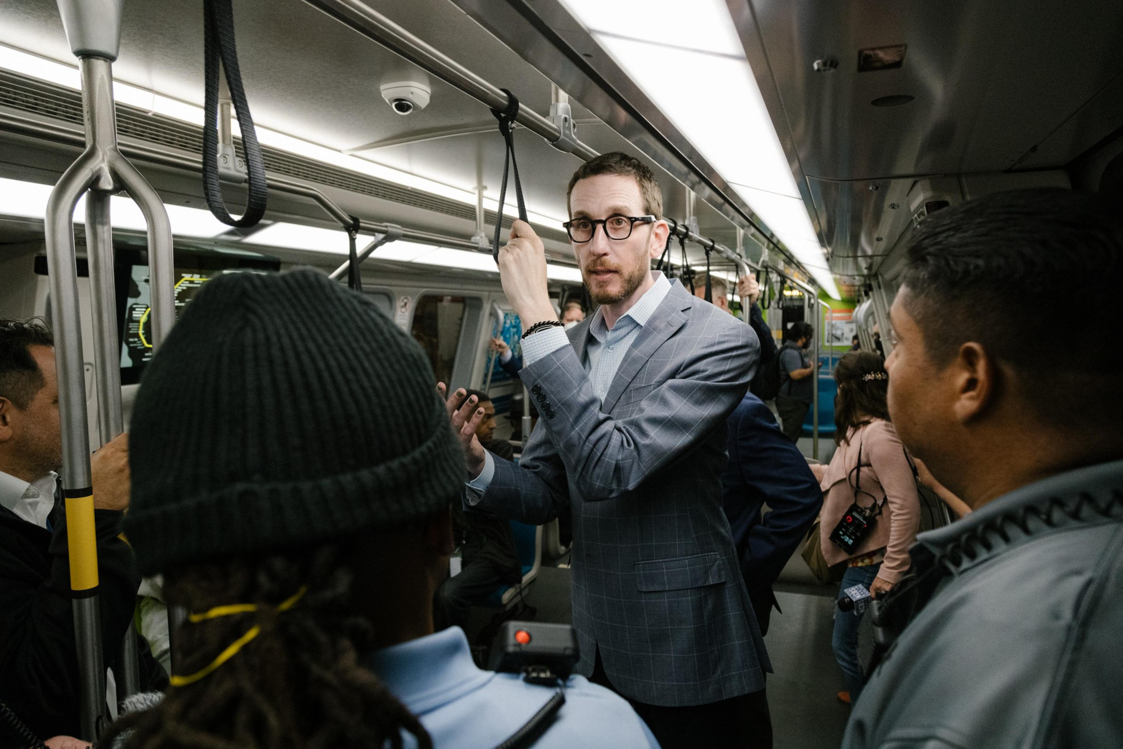 A man in a gray suit and classes stands with his hand in a strap on a train.