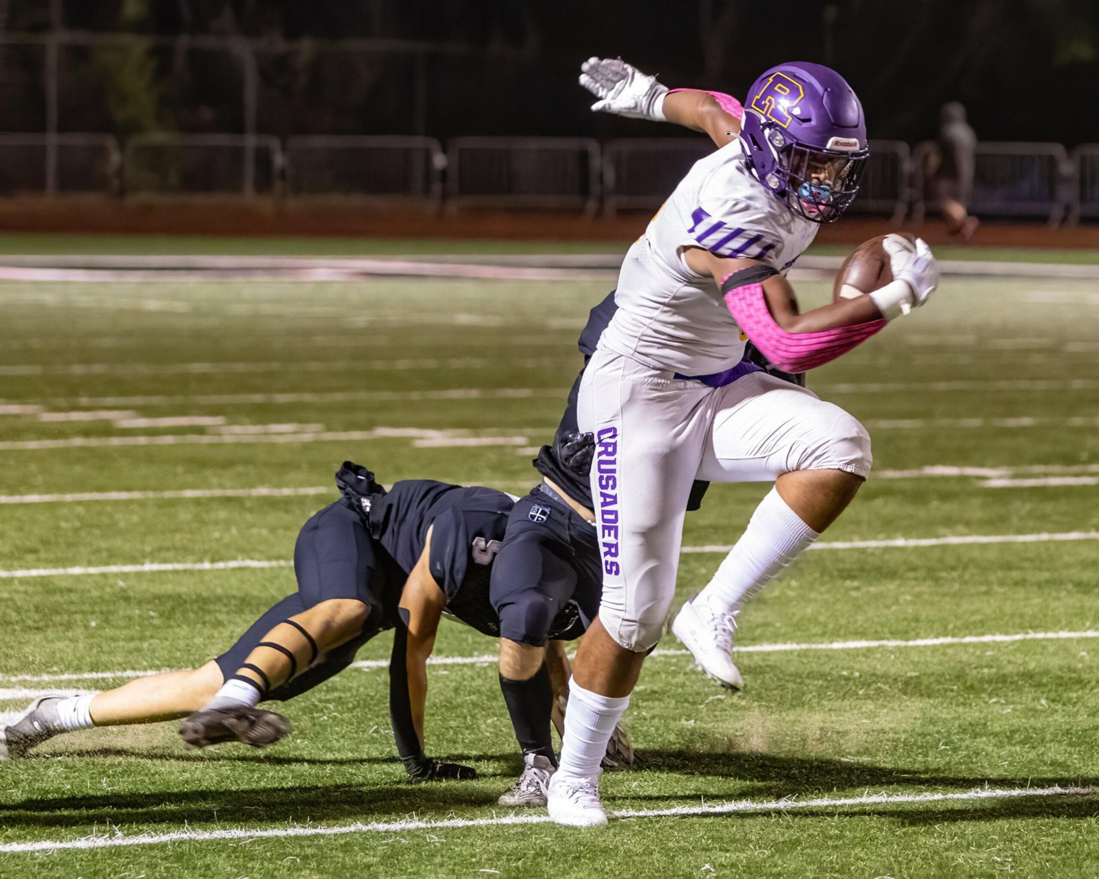 A football player in a white uniform with "Crusaders" on his pants runs with the ball while a player in black falls behind him on the field.