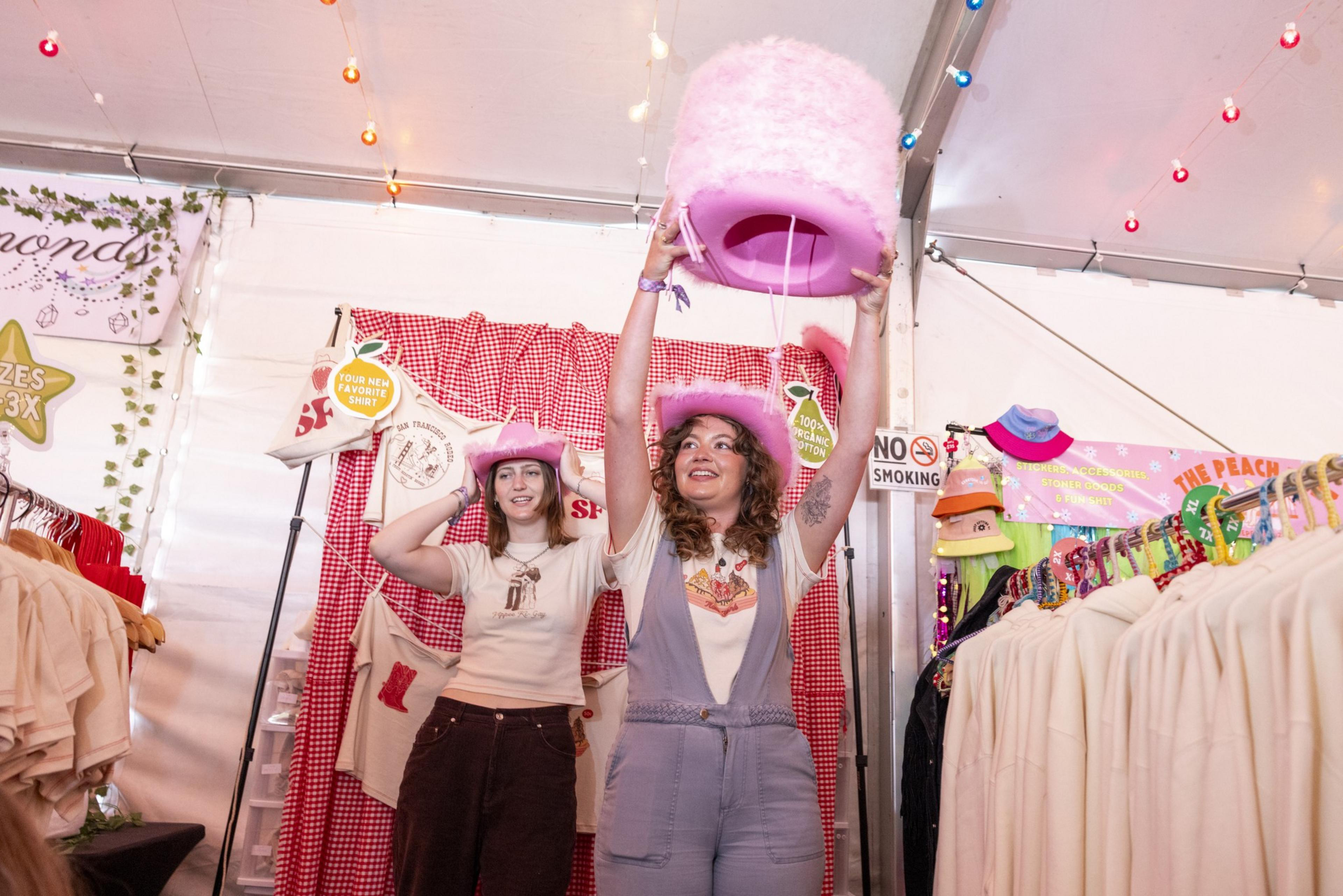 Two people are inside a vibrant, decorated booth. They are smiling and wearing pink cowboy hats, with one person lifting a large pink fluffy hat above their head.