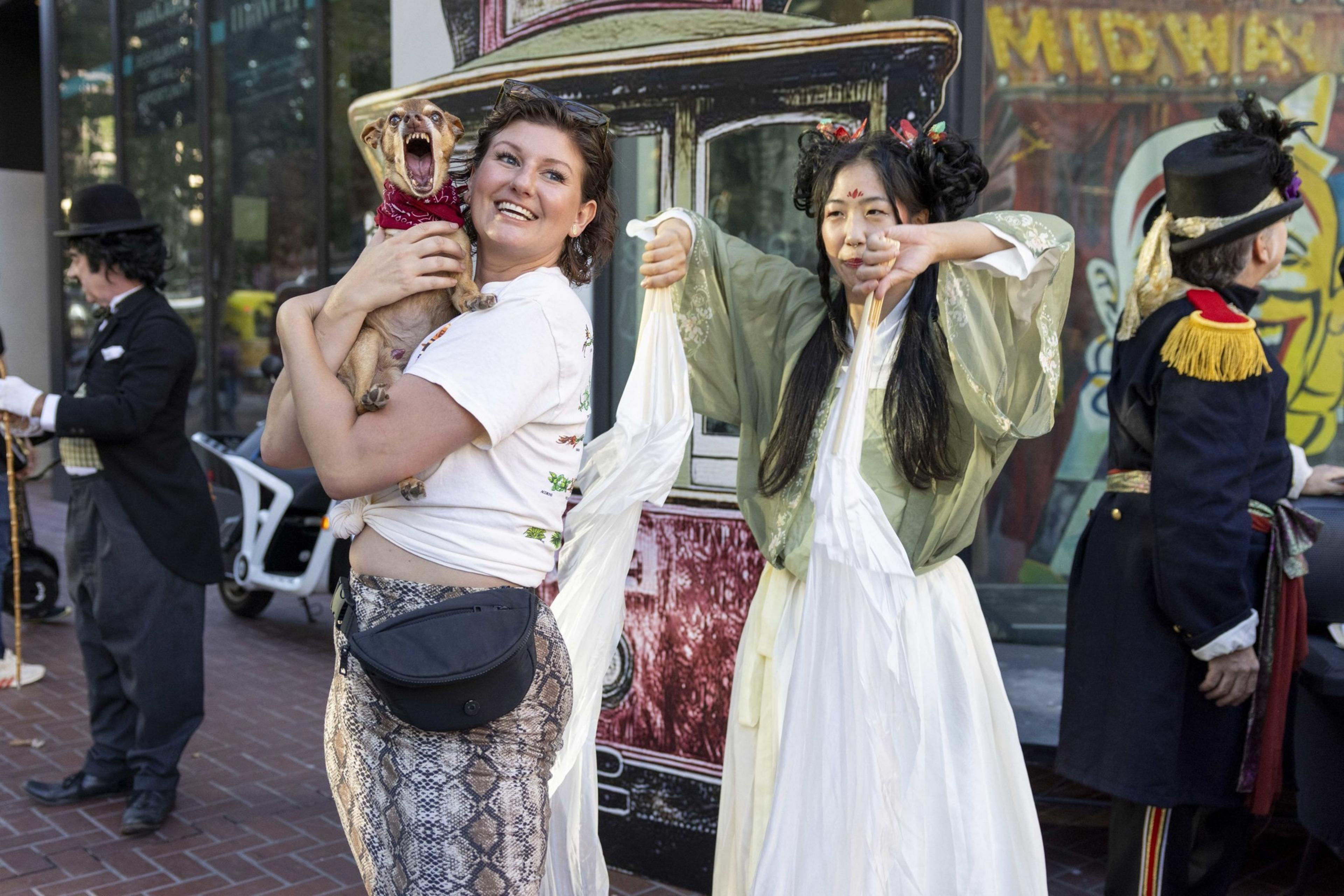 A smiling woman holds a small dog while another woman in traditional costume dances; people in vintage attire are seen in the background.