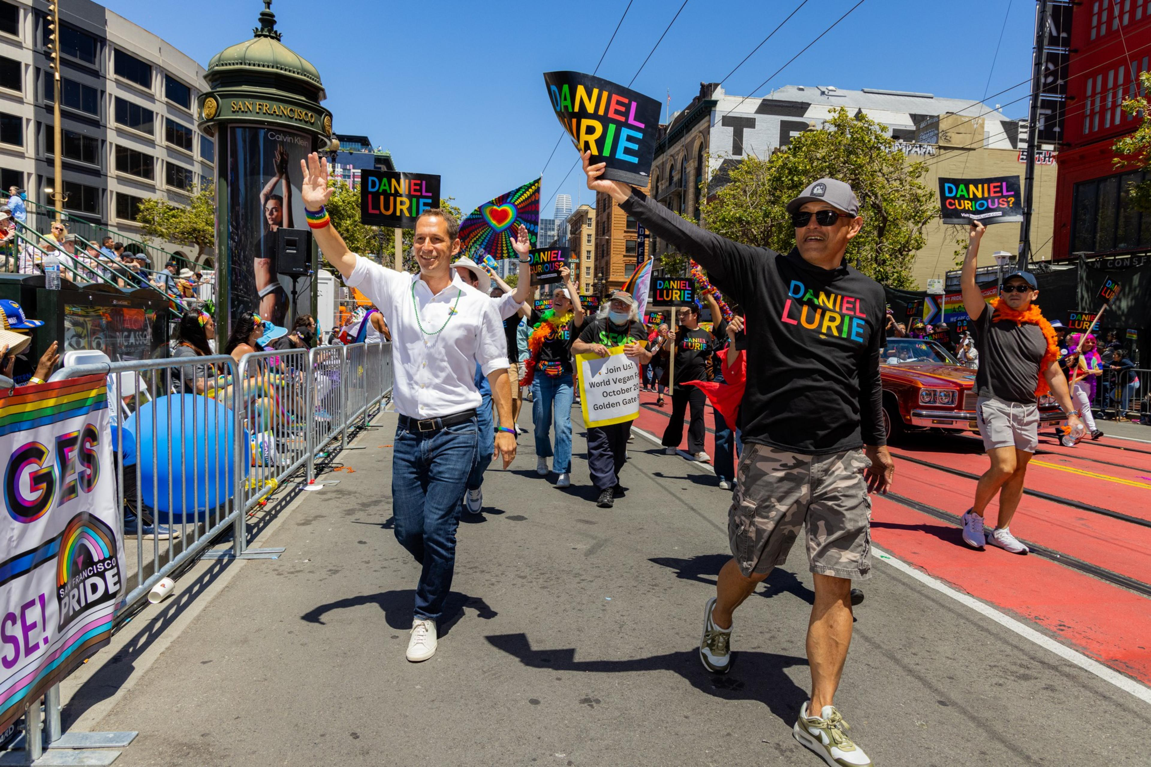 People parade in a vibrant street celebration, holding colorful &quot;Daniel Lurie&quot; signs, with spectators and rainbow flags behind barricades. The atmosphere is festive and joyful.