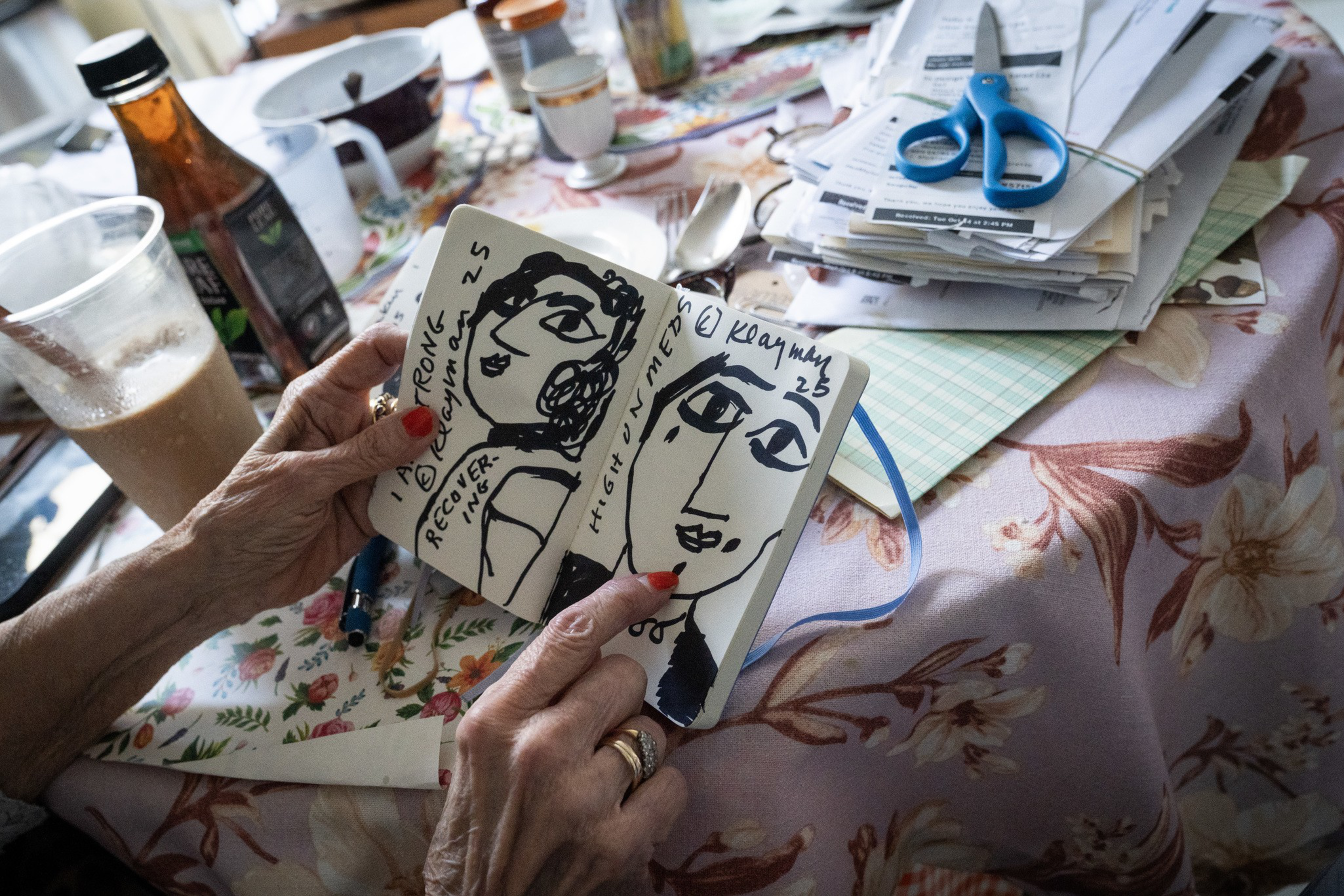 An elderly person with red-painted nails holds an open sketchbook showing two abstract black ink portraits with handwritten notes on each page.