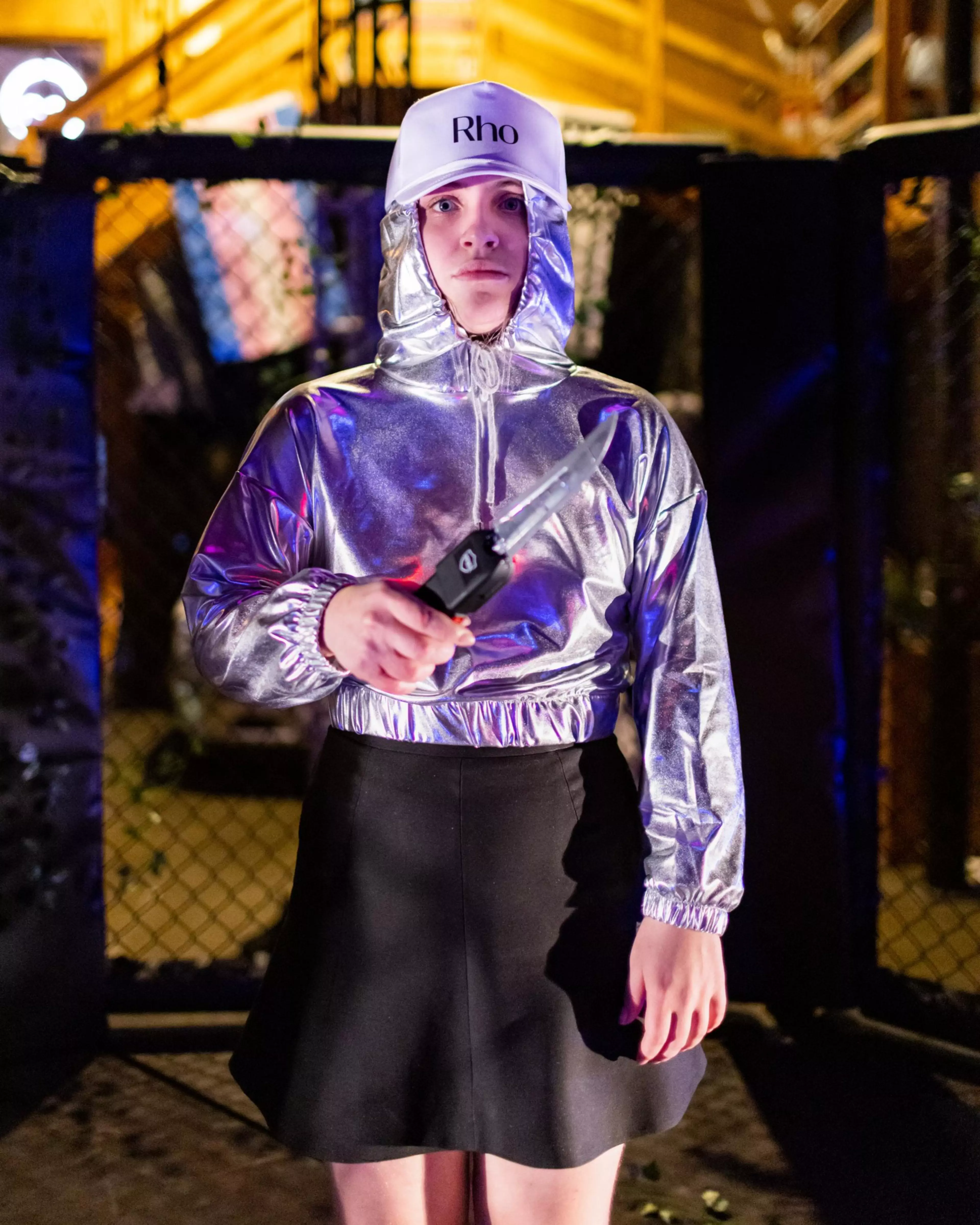 A person wearing a shiny silver hooded top, black skirt, and white cap labeled “Rho” holds a large folding knife, standing in front of a dark fence.