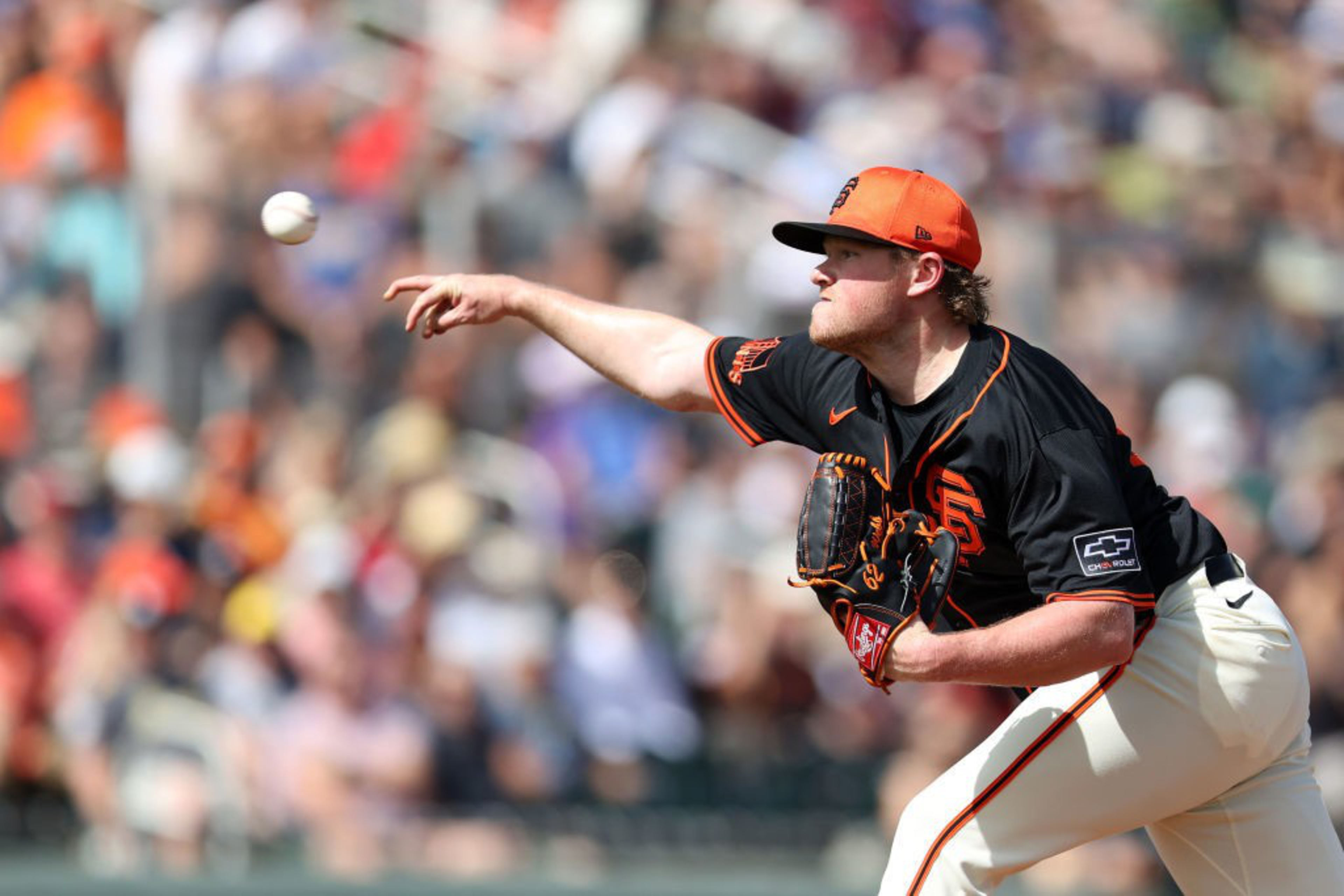 A baseball player in a black and orange uniform pitches the ball, mid-action, with a blurred crowd in the background.