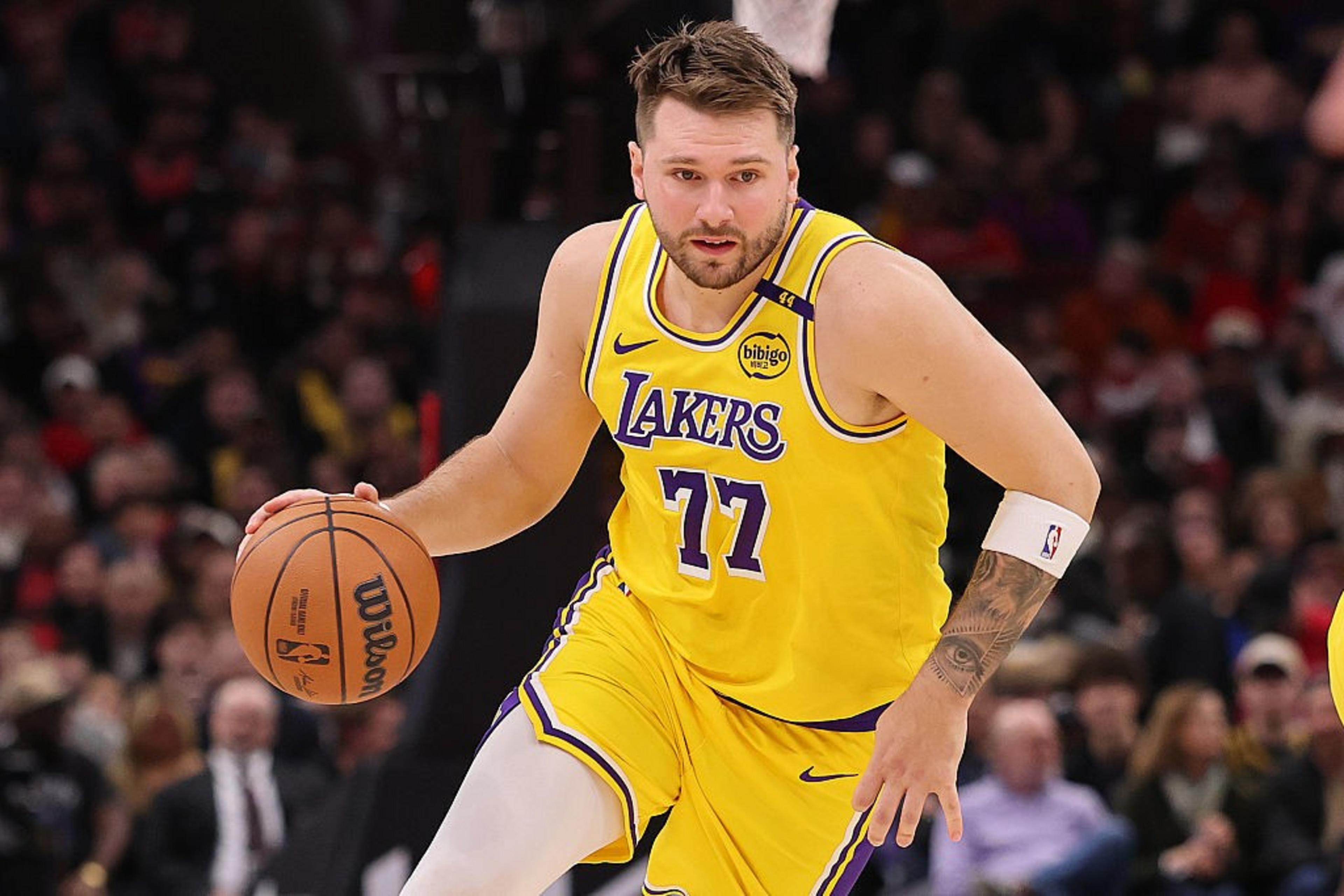 A basketball player in a yellow Lakers jersey, number 77, dribbles the ball on a court. The background is blurry with spectators watching the game.
