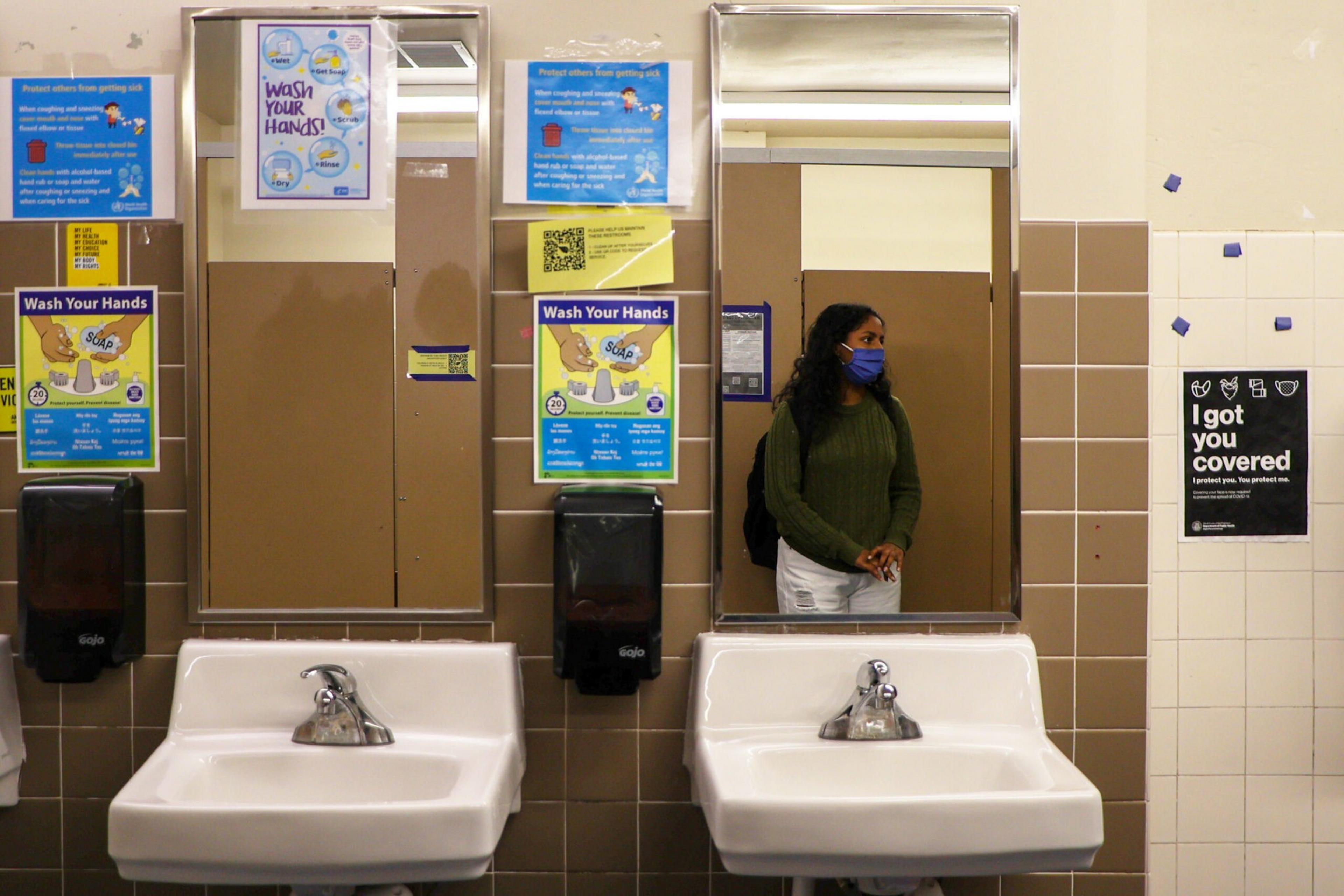A woman wearing a blue mask and green sweater stands in front of two sinks and mirrors in a restroom with several signs about washing hands and staying covered.