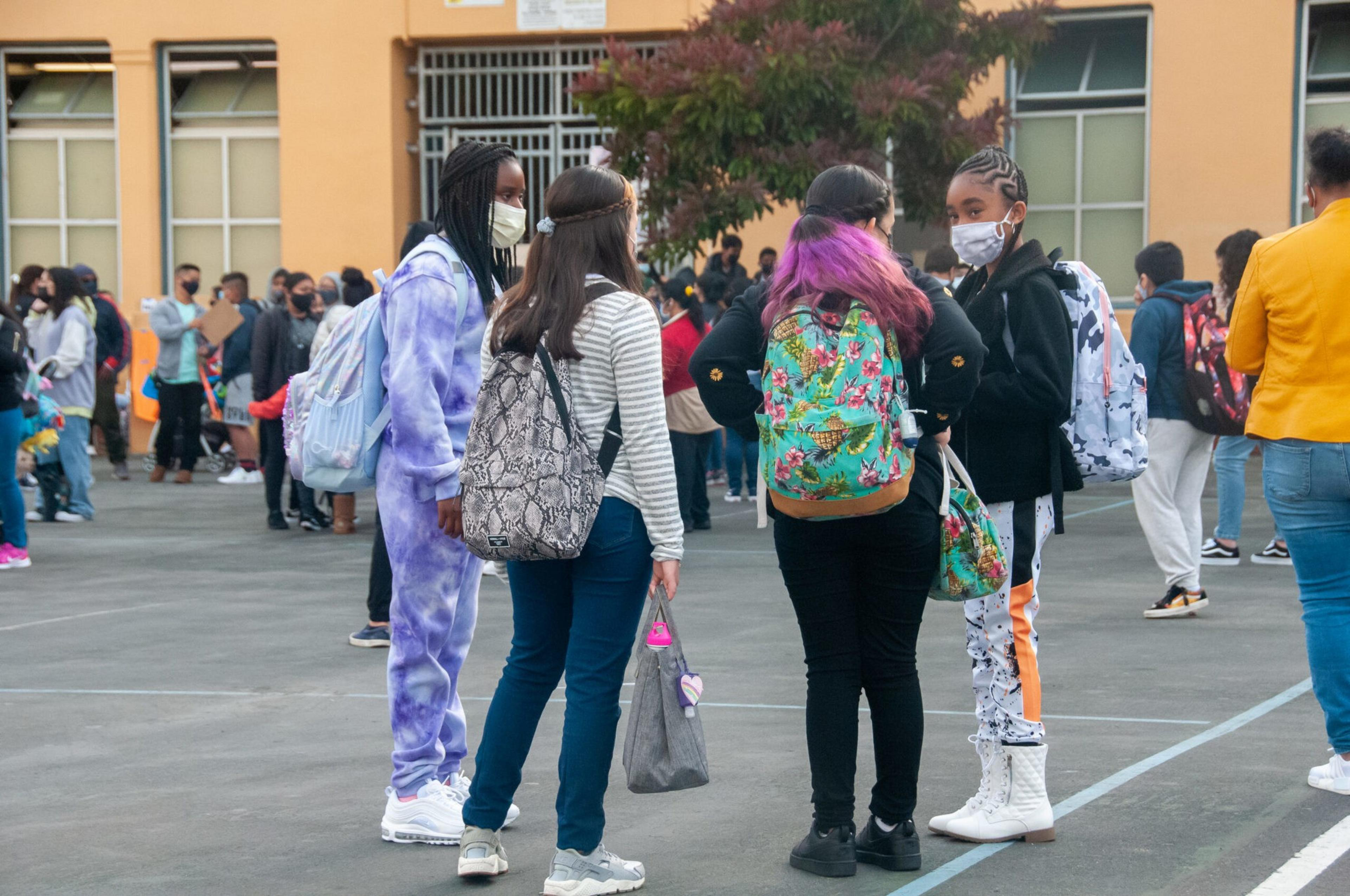 A group of students wearing backpacks and face masks stand talking in a school courtyard, with more students and a building in the background.