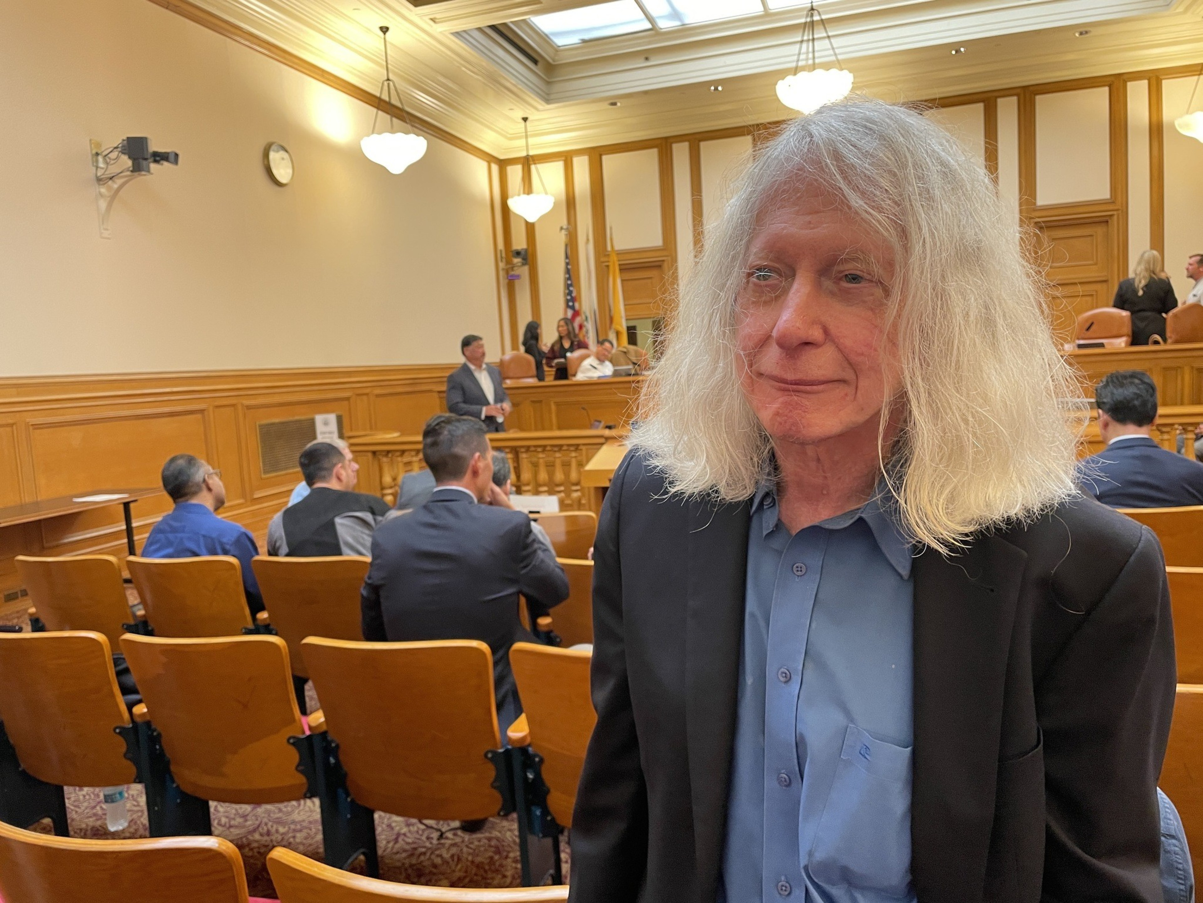 A man with long white hair, wearing a black jacket and blue shirt, stands in the foreground of a courtroom filled with people seated and someone speaking up front.