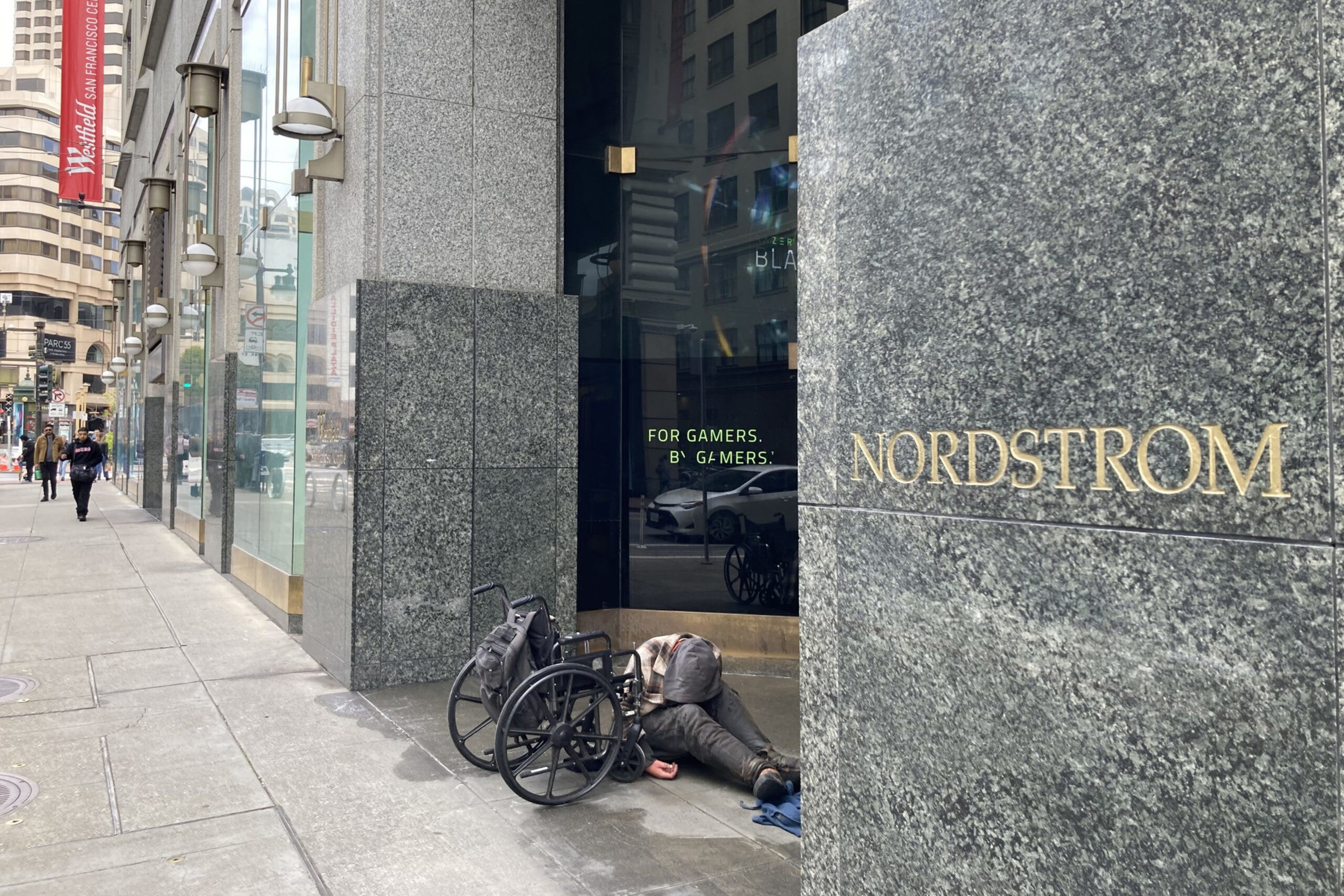 A person lies on the ground beside an empty wheelchair outside a Nordstrom store in a city setting.