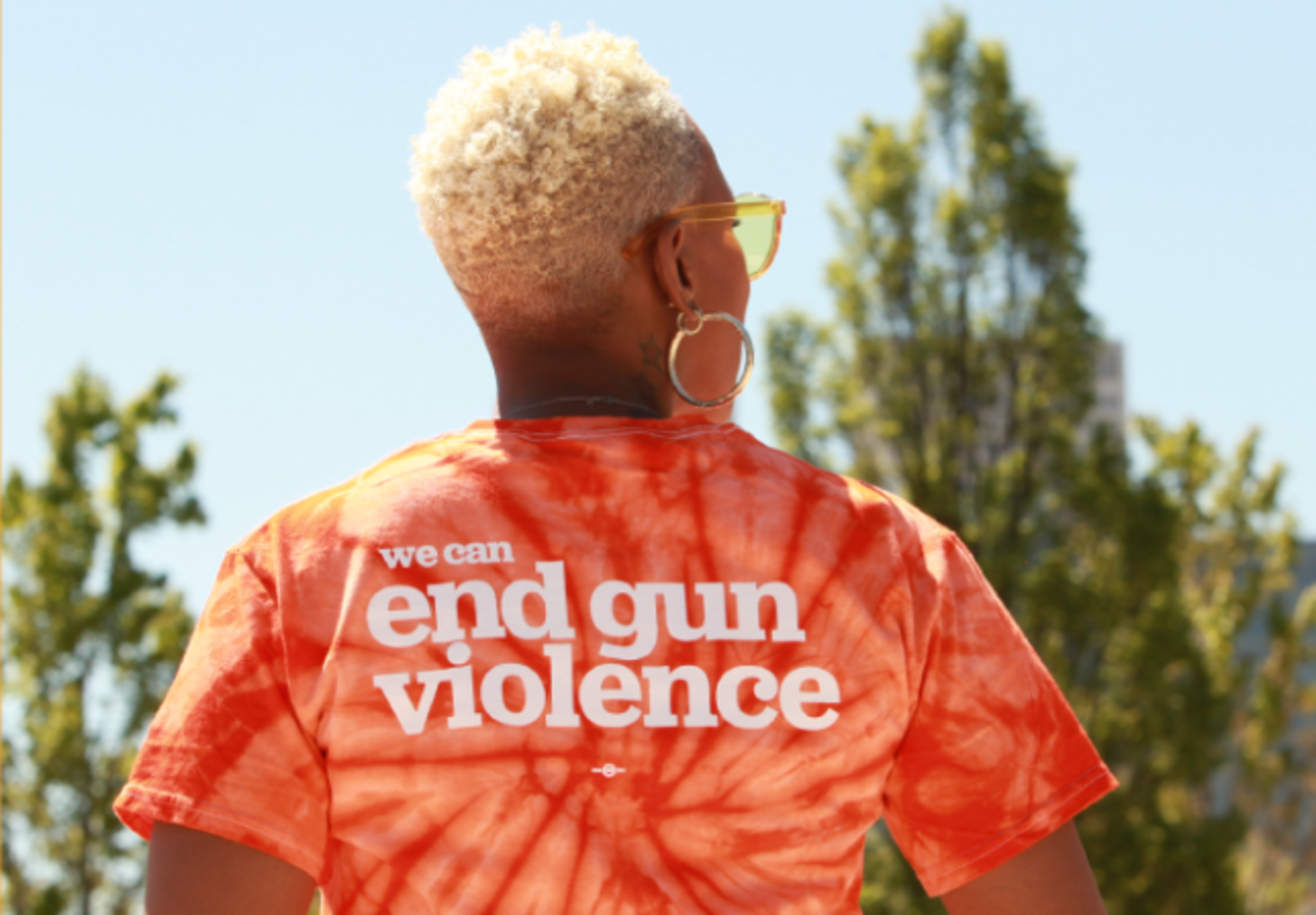 A person with short blonde hair wears an orange tie-dye shirt that reads "we can end gun violence" in white letters, outdoors with trees behind them.