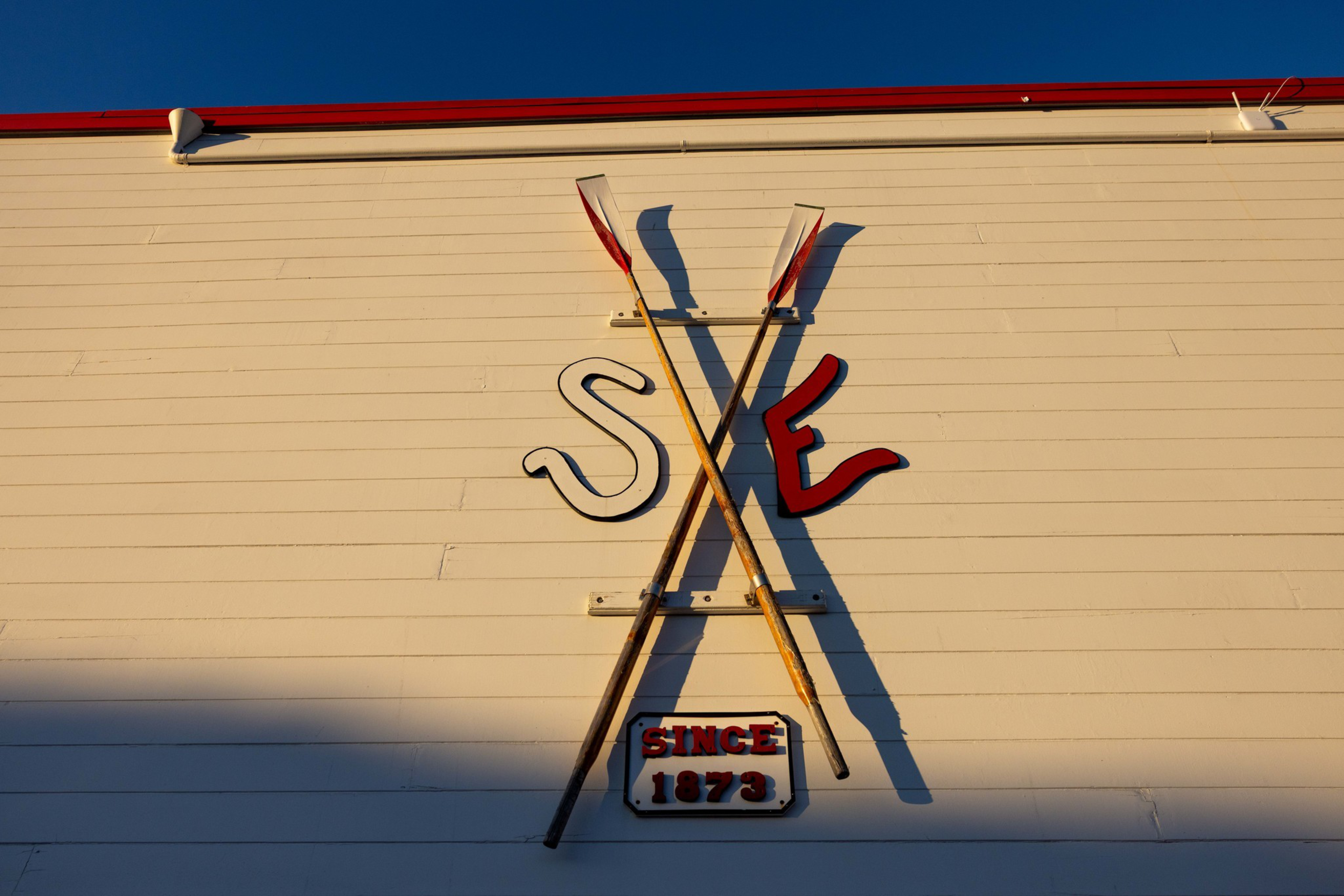 Two crossed wooden oars hang on a cream-colored wall with red letters “S” and “E” on either side and a sign below reading “Since 1873.”