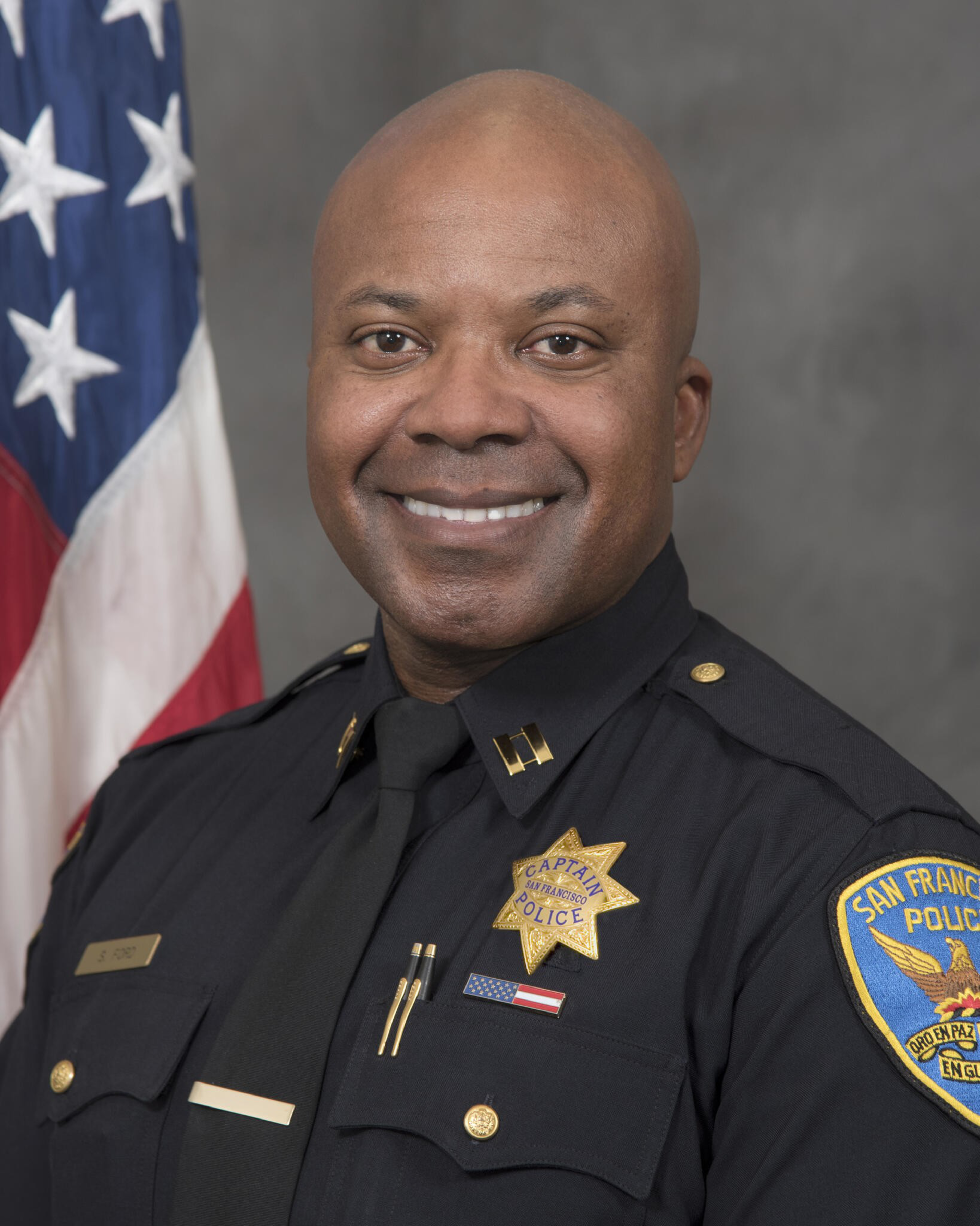 A smiling African American police captain in a San Francisco Police uniform stands in front of a U.S. flag and gray background.