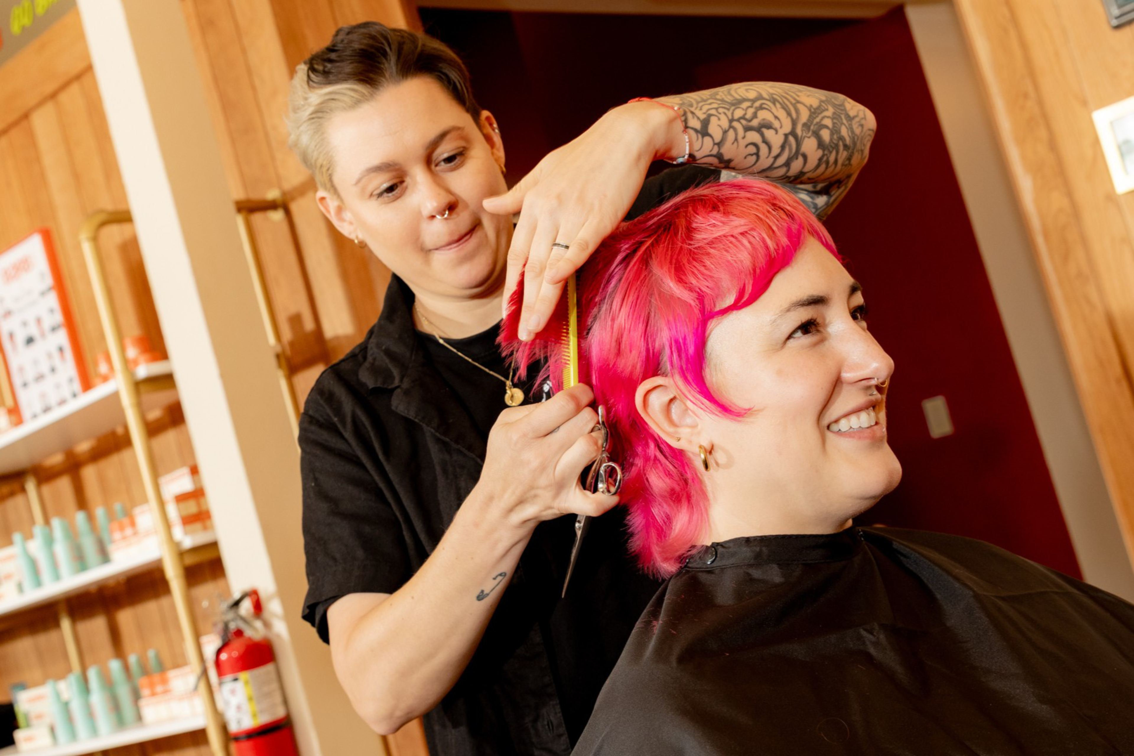 A hairstylist with tattooed arms trims the bright pink hair of a smiling client seated in a salon.
