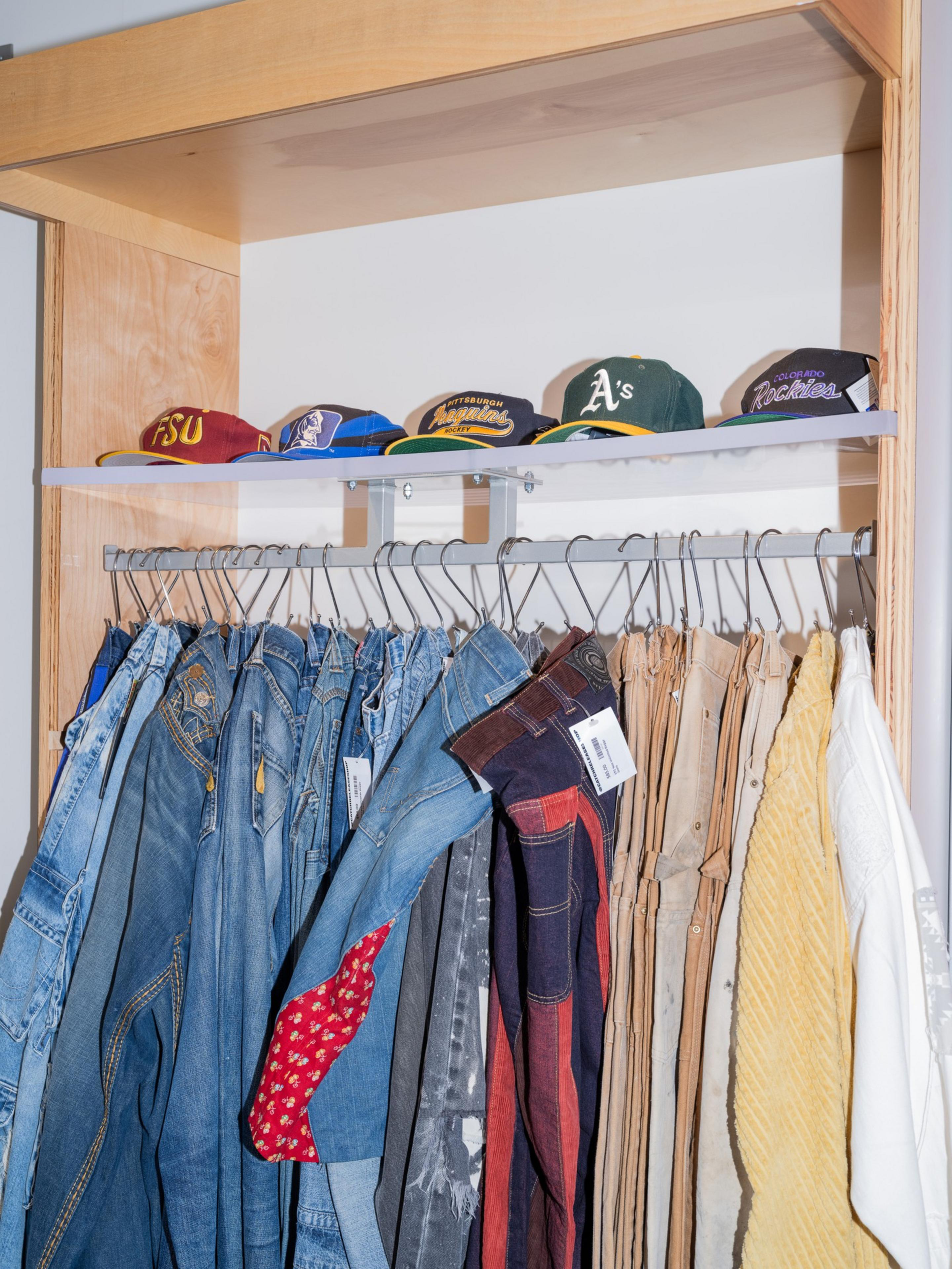 The image shows a row of colorful hats on a shelf above a rack of jeans and shirts hanging on hangers, with labels attached to the clothes.