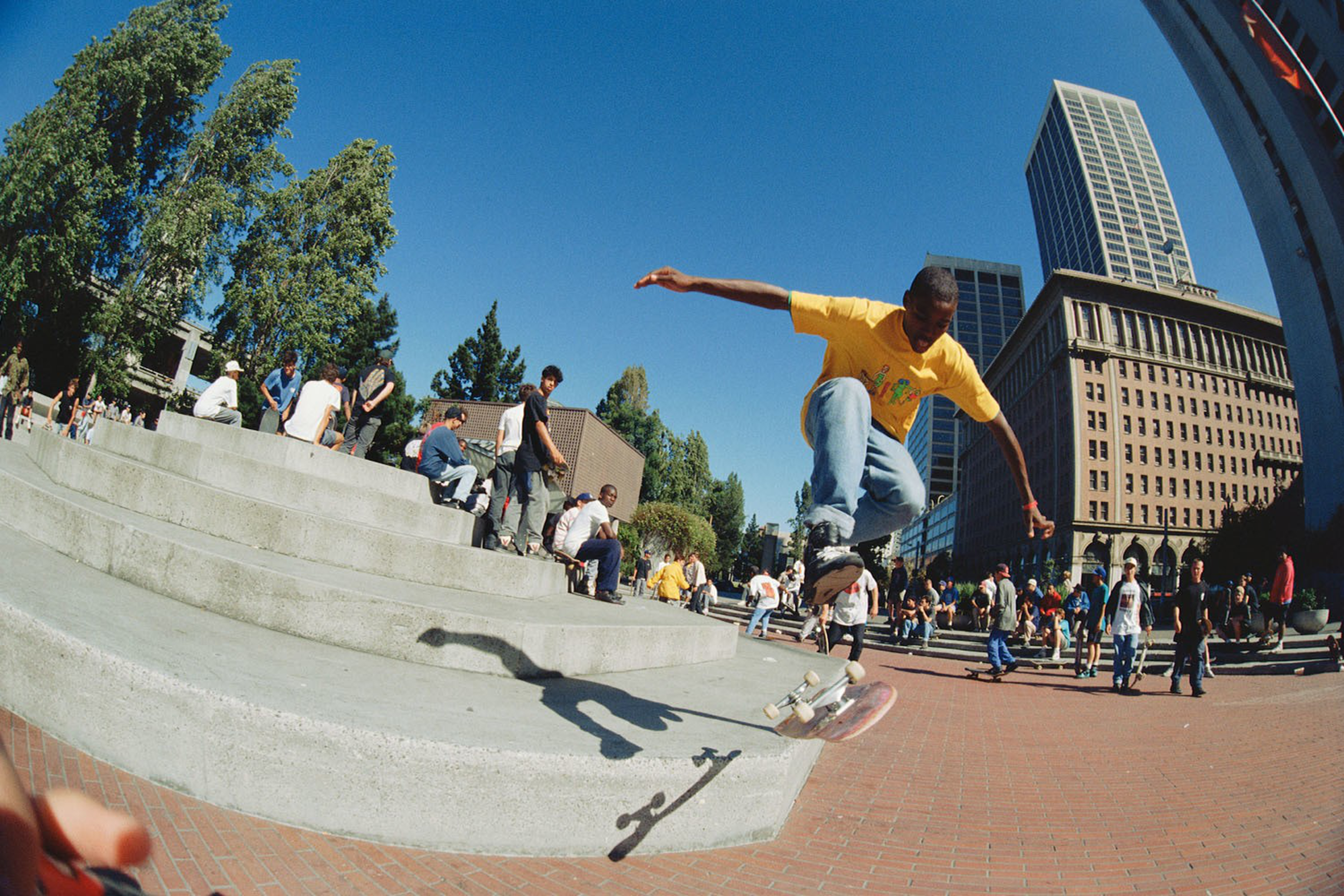 A young man in a yellow shirt performs a skateboard trick off concrete steps while a crowd watches in an urban plaza with tall buildings and trees.