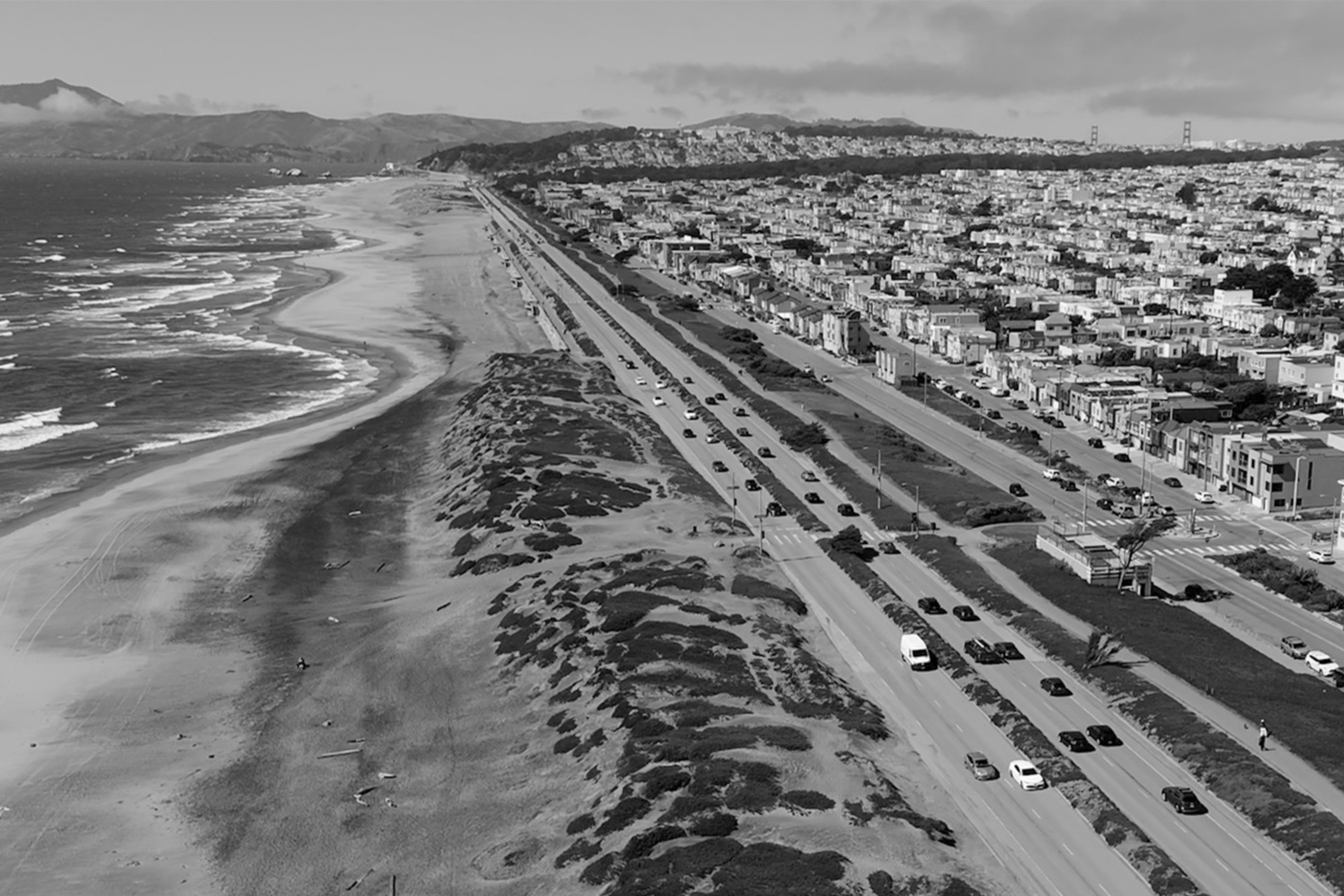 A coastal scene shows a long beach with waves on the left and a busy road bordered by houses on the right. The area is bustling with traffic.