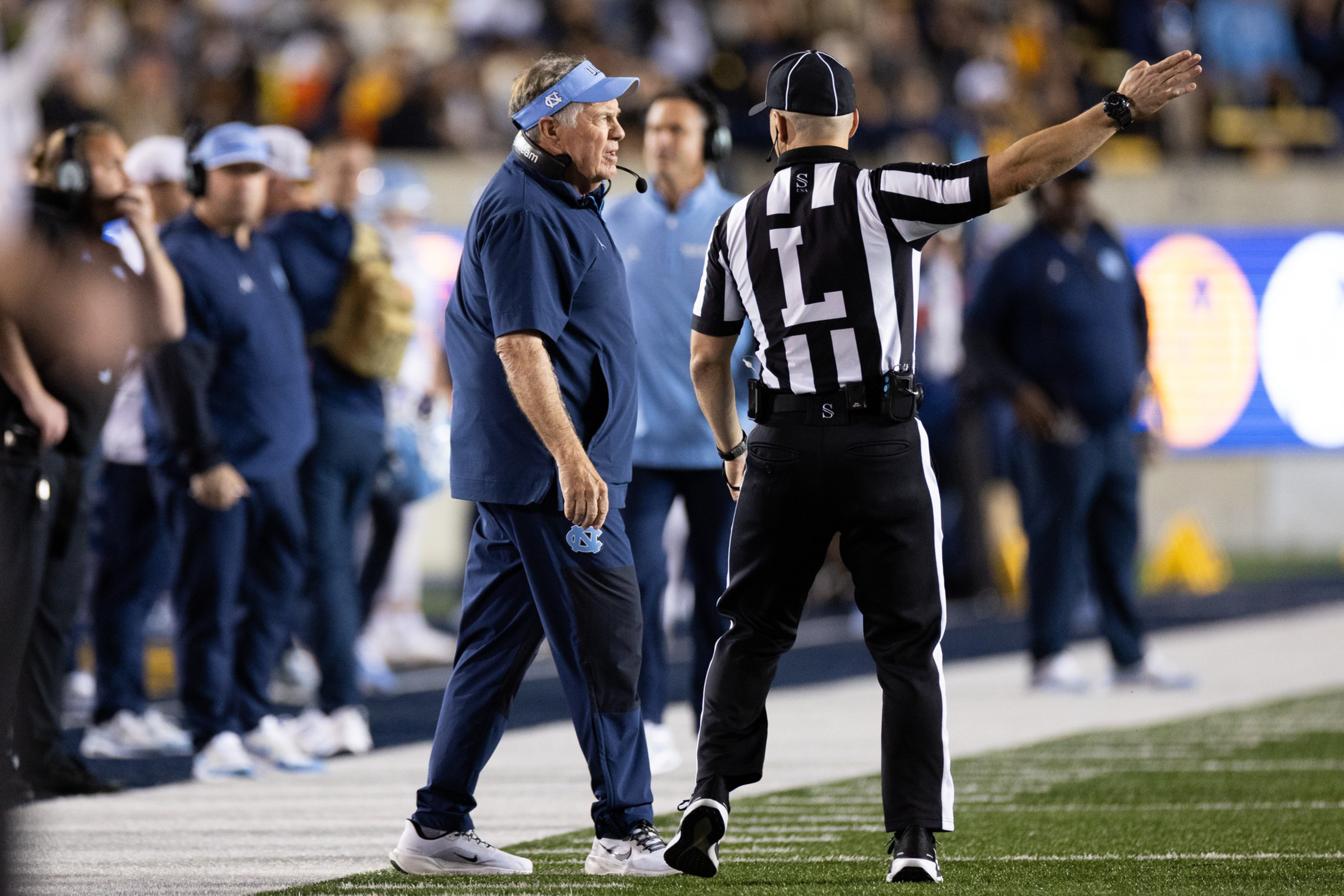 A football coach in navy blue argues with a referee in black-and-white stripes on the sideline during a game.
