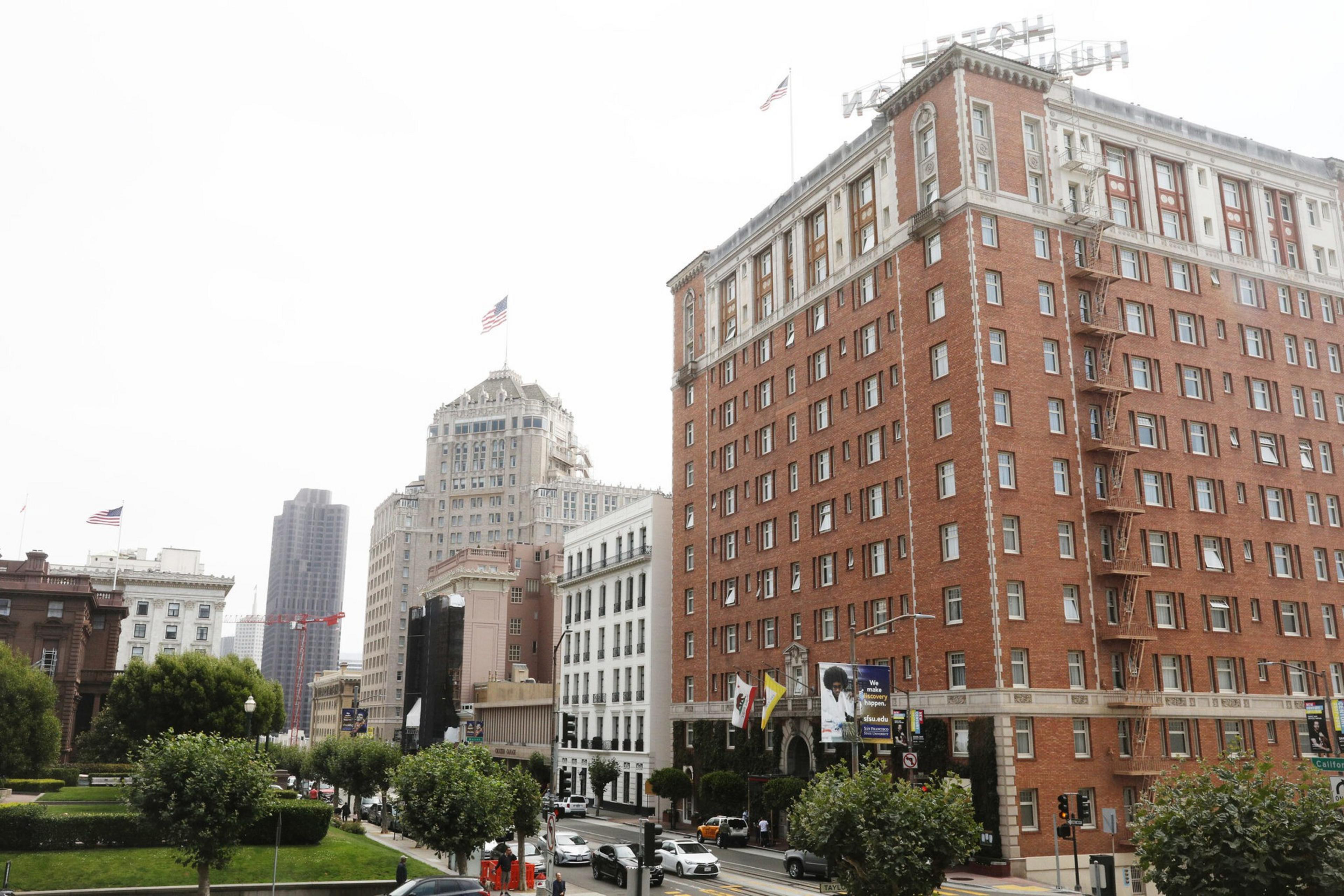 A busy urban street bordered by trees and large, multi-story historic buildings, including a prominent brick hotel with fire escapes and flags.