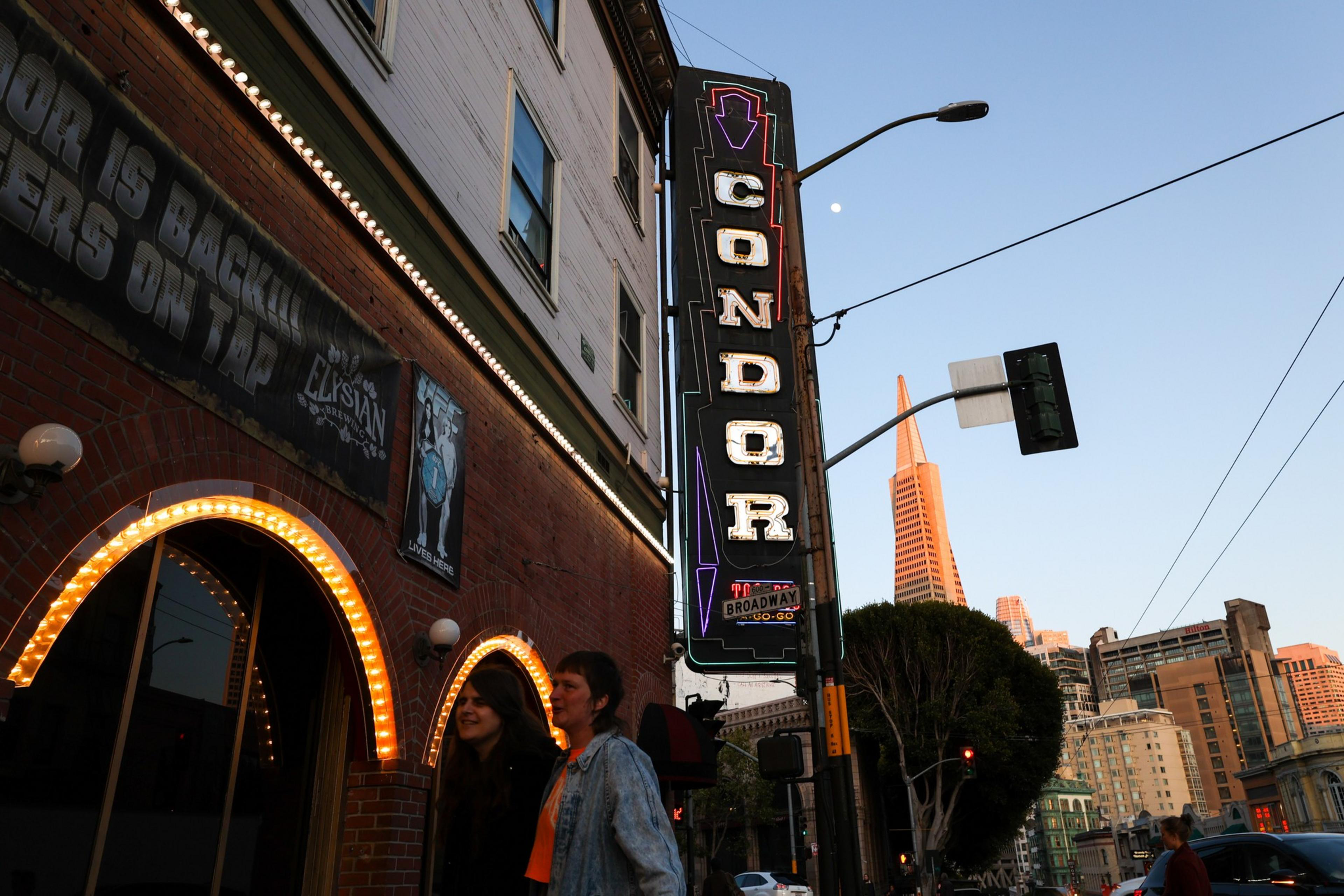 A lit "CONDOR" sign hangs on a building with two women standing below, and a city skyline including a pointed skyscraper is visible in the background.