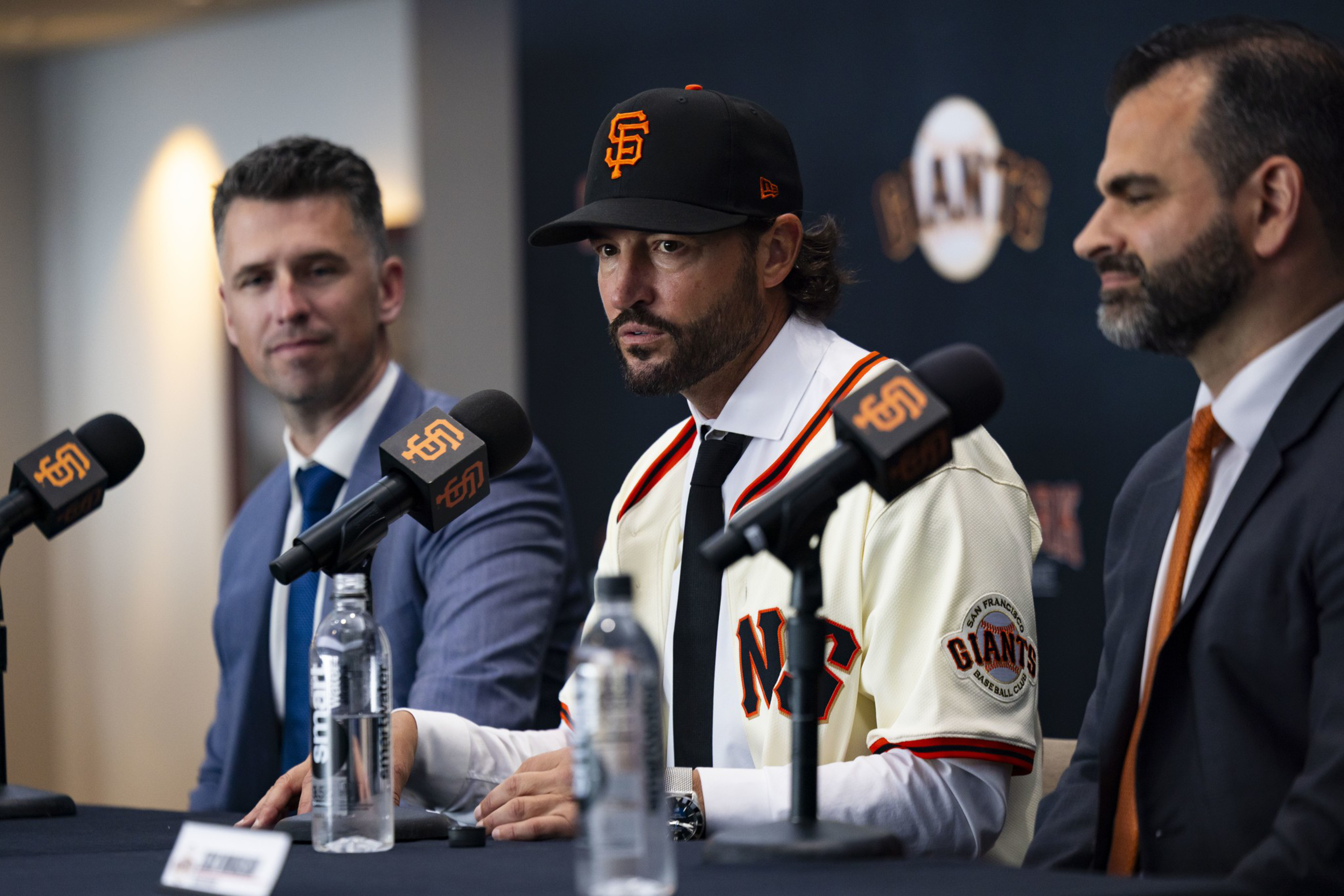 Three men sit at a table with microphones; the center man wears a San Francisco Giants baseball cap and jersey, flanked by two men in suits.