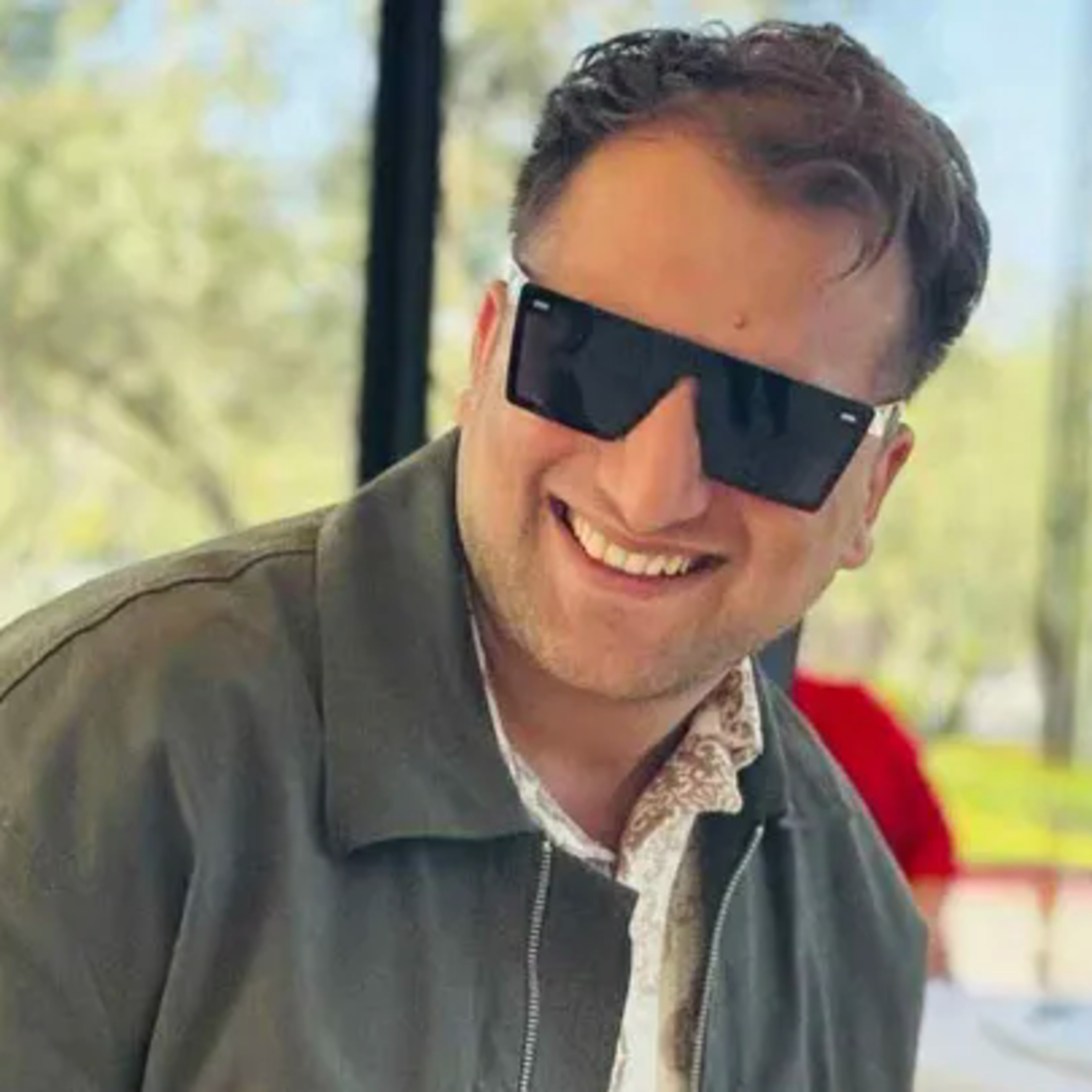 A man smiles broadly while wearing large black sunglasses and a green jacket, with a blurred outdoor background featuring trees and another person.